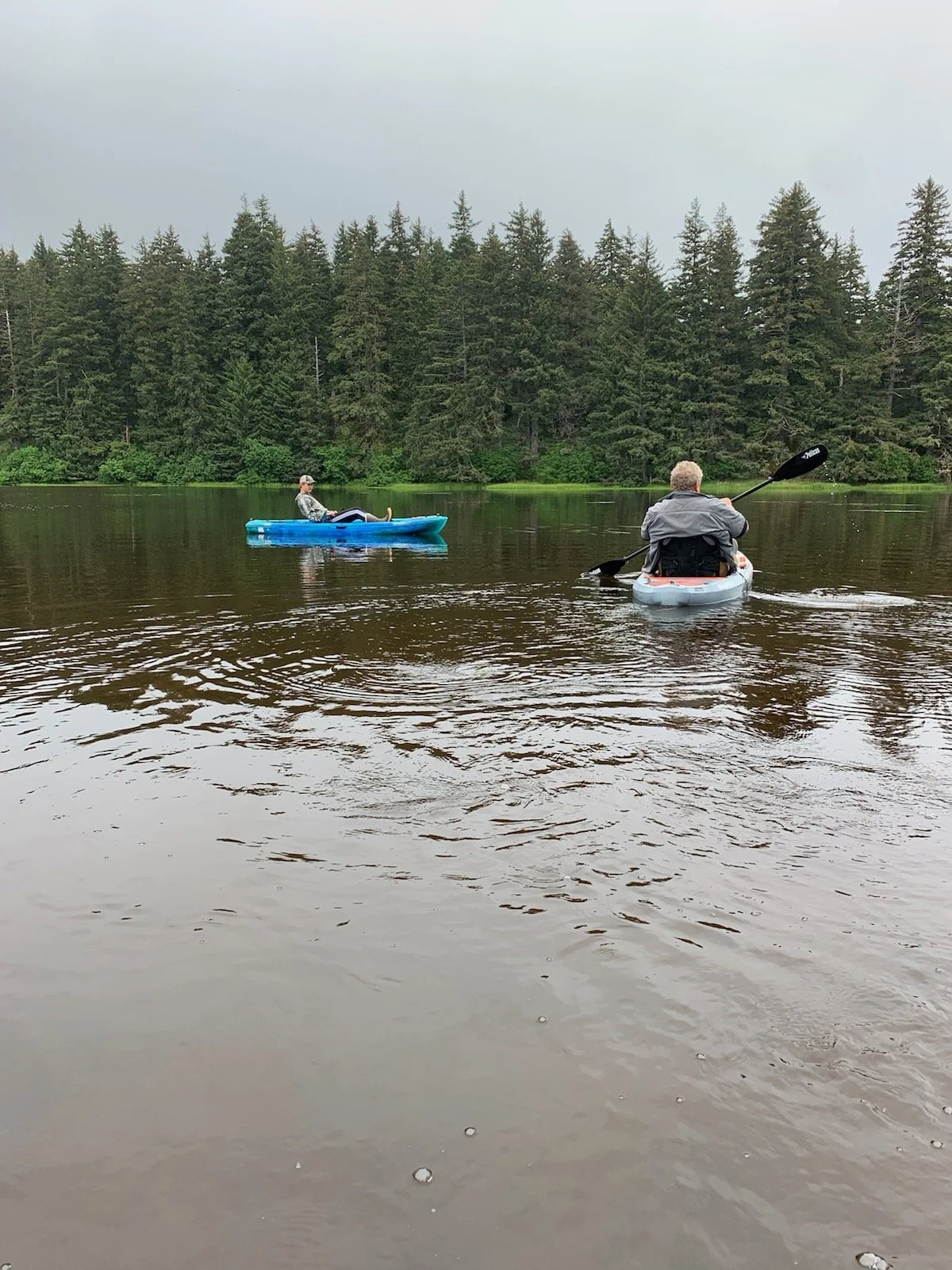 Two people kayaking on a calm lake with a forested shoreline in the background.