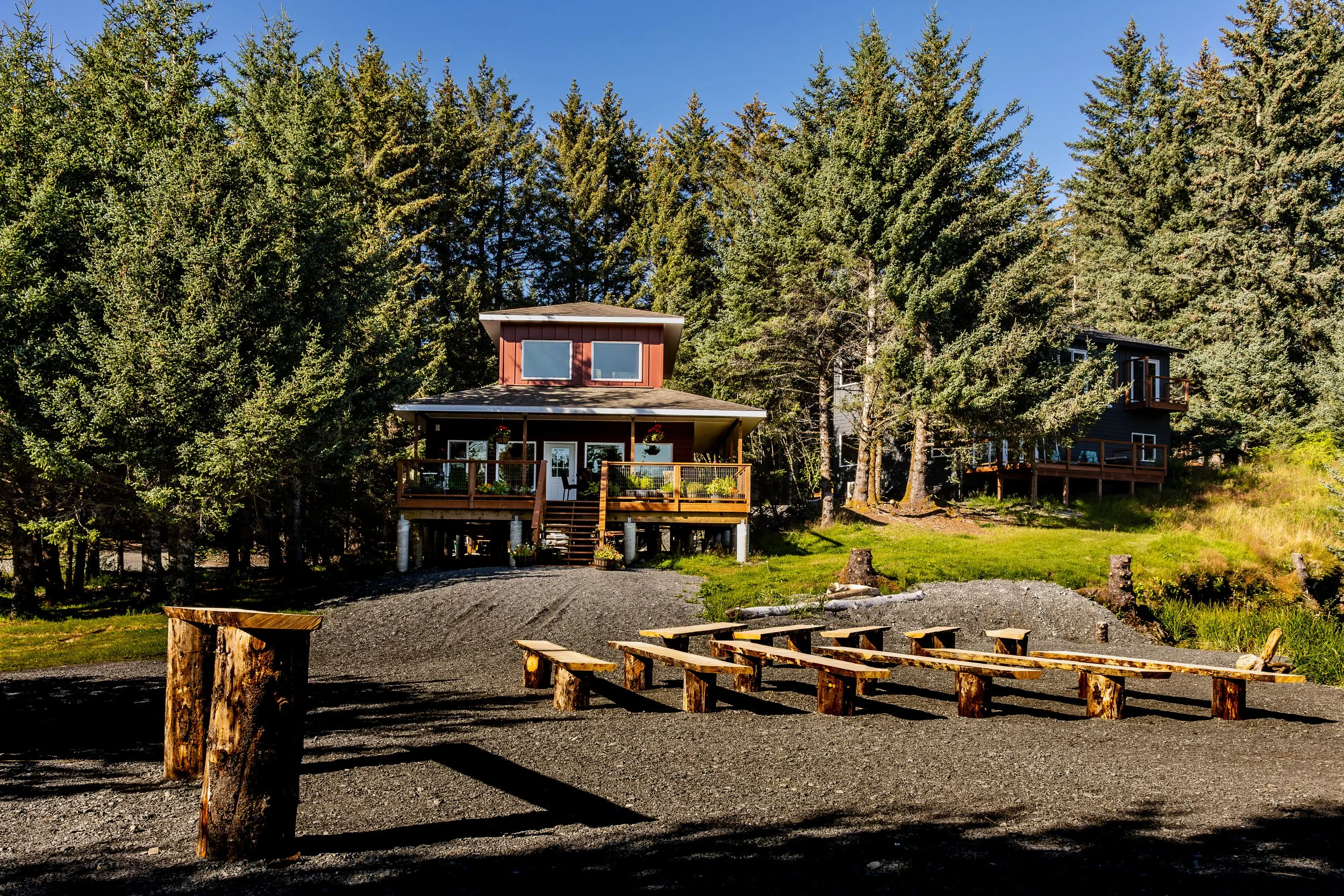 A cottage built on the edge of a forest with tall evergreen trees, a spacious front porch with string lights, and a deck with a small balcony. There are outdoor benches made from logs and a gravel ground area with a row of logs as benches.