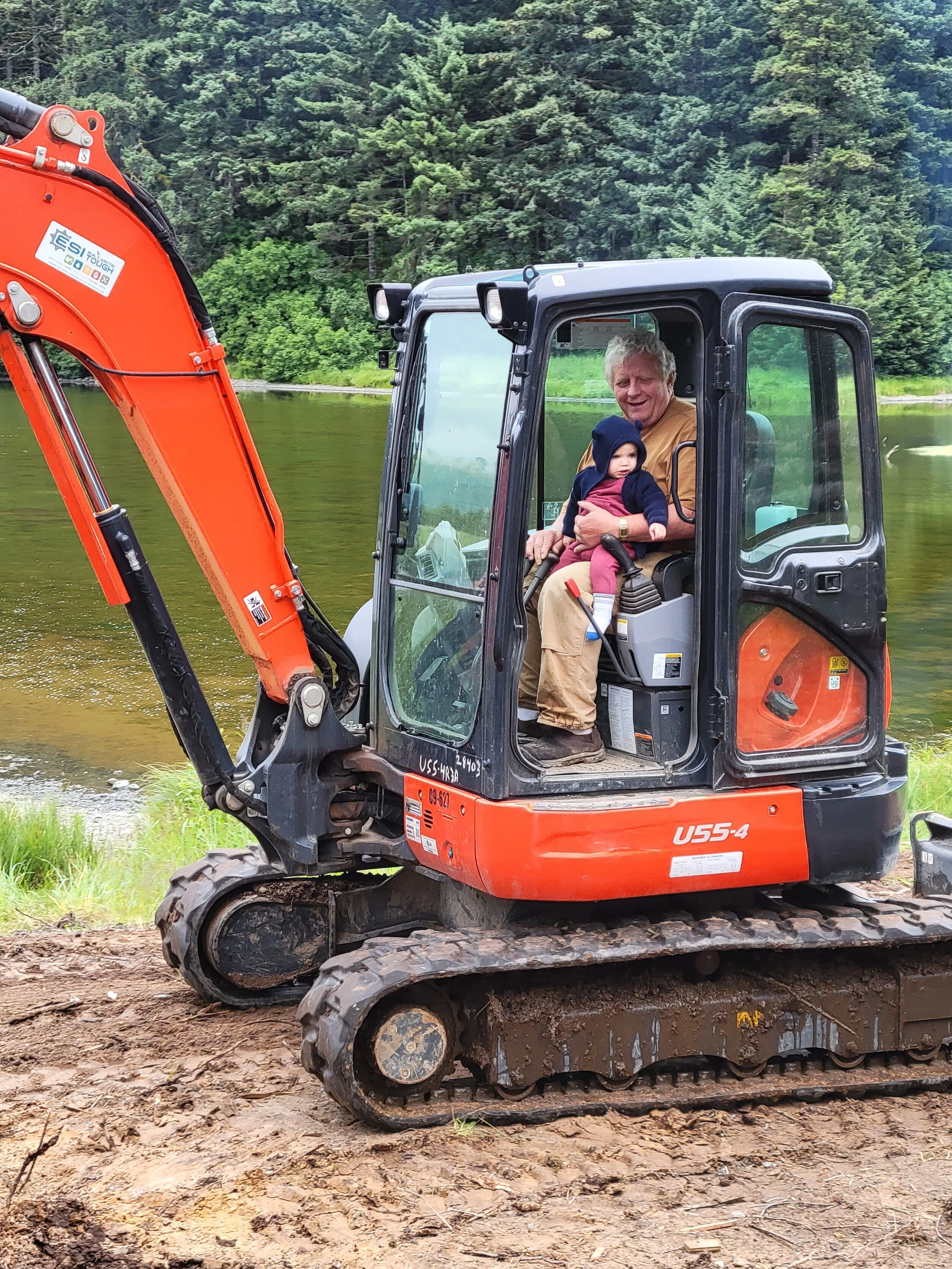 A man and a baby sitting inside a small orange and black mini excavator machine near a forested area with a body of water behind them.