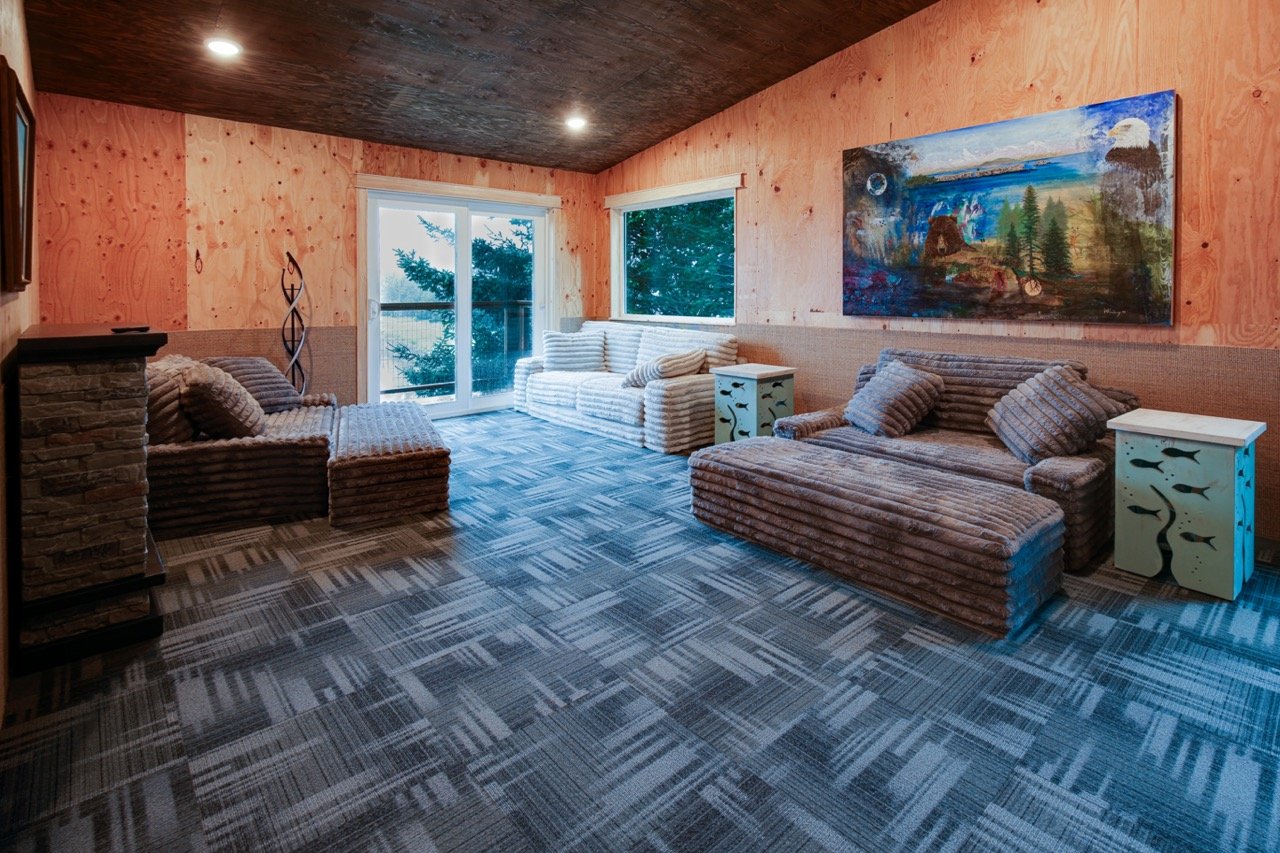 Living room with plaid sofas, striped cushions, wooden walls, mural painting, and windows showing trees outside.