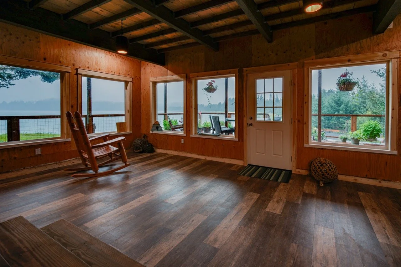 Interior of a wooden room with large windows, a rocking chair, hanging flower baskets, potted plants on the windowsills, and a view of a lake and trees outside.