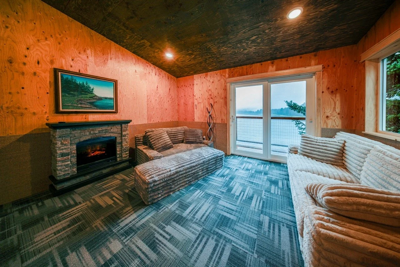 Living room with wood-paneled walls and ceiling, a fireplace, two sofas, a sliding glass door leading to a balcony with a lake view, and a painting above the fireplace.