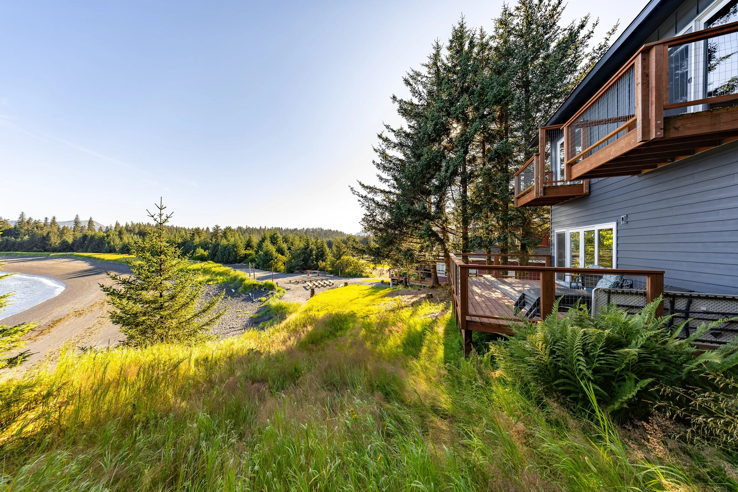 A cottage with wooden balconies overlooking a grassy area with trees and a beach in the background under a clear sky.