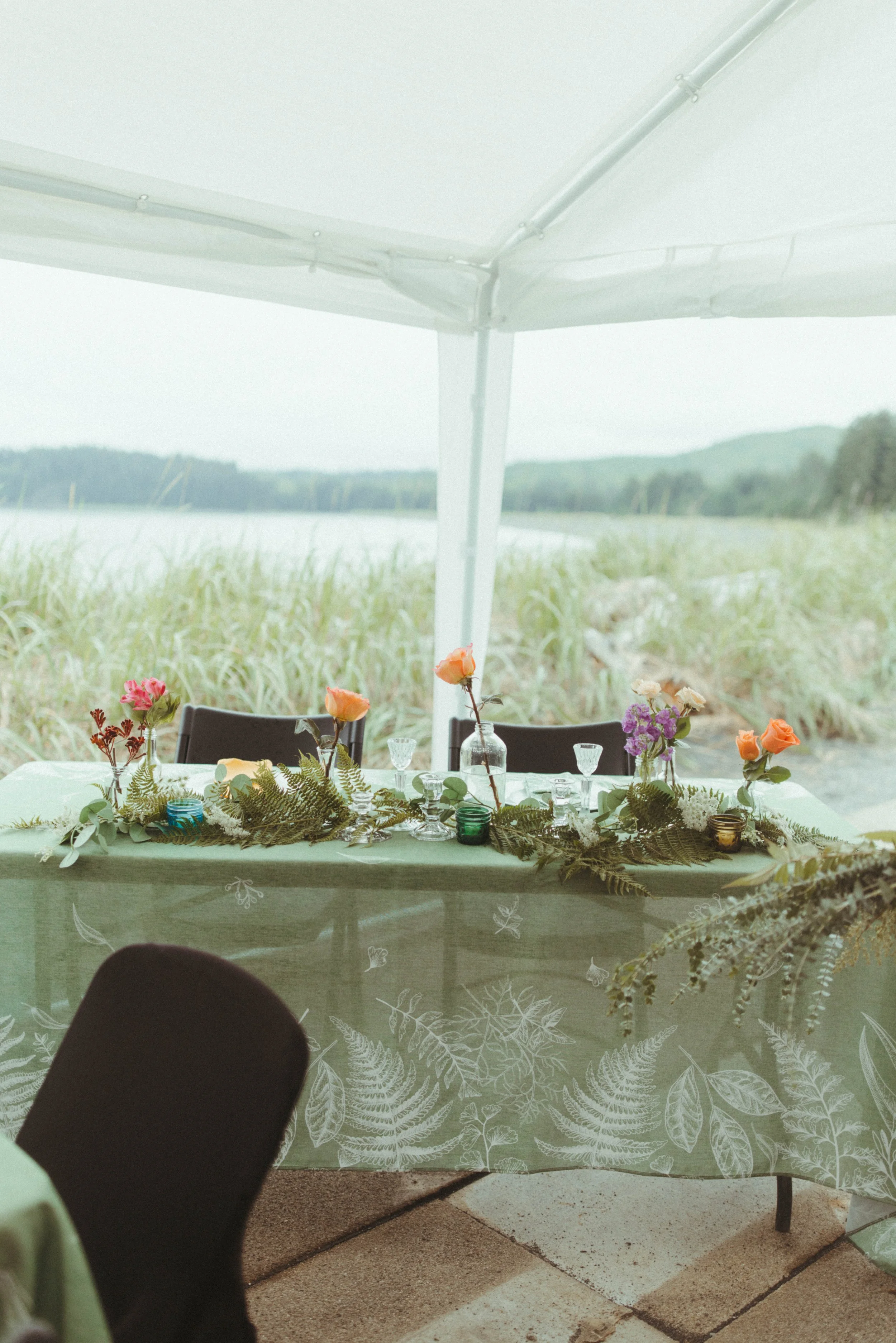 Outdoor dining table decorated with flowers and greenery, set under a large white canopy overlooking a grassy area and distant hills.
