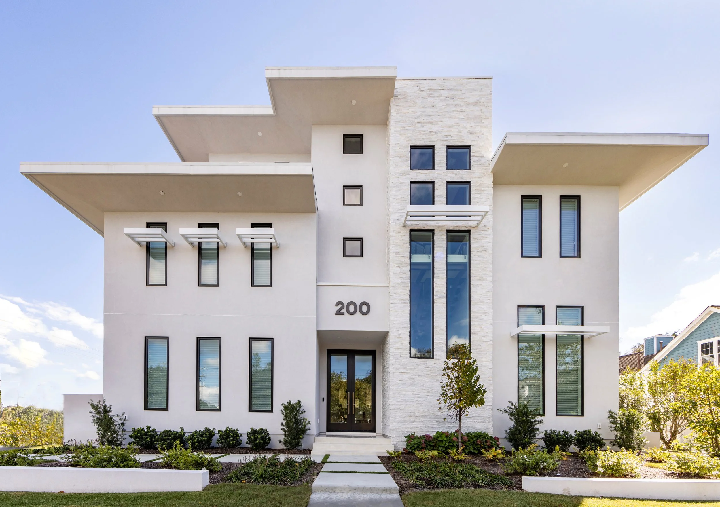 Modern white multi-story house with large windows, flat overhanging roofs, and landscaped front yard.