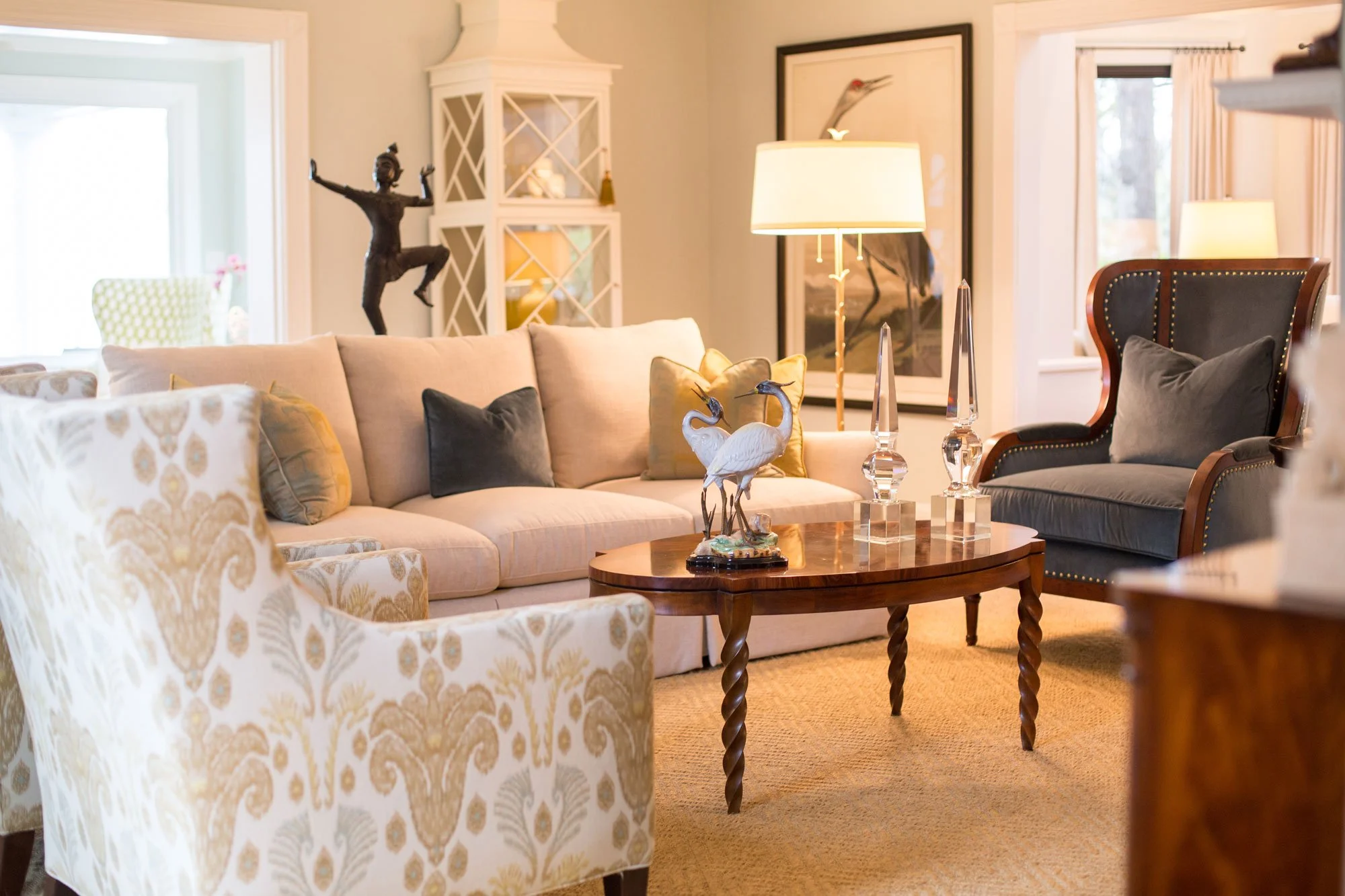 Living room with beige sofa, patterned armchairs, dark armchair, wooden coffee table decorated with swan sculpture and glass decorative pieces, floor lamp, framed animal print, and sculptures on a white cabinet near a window.