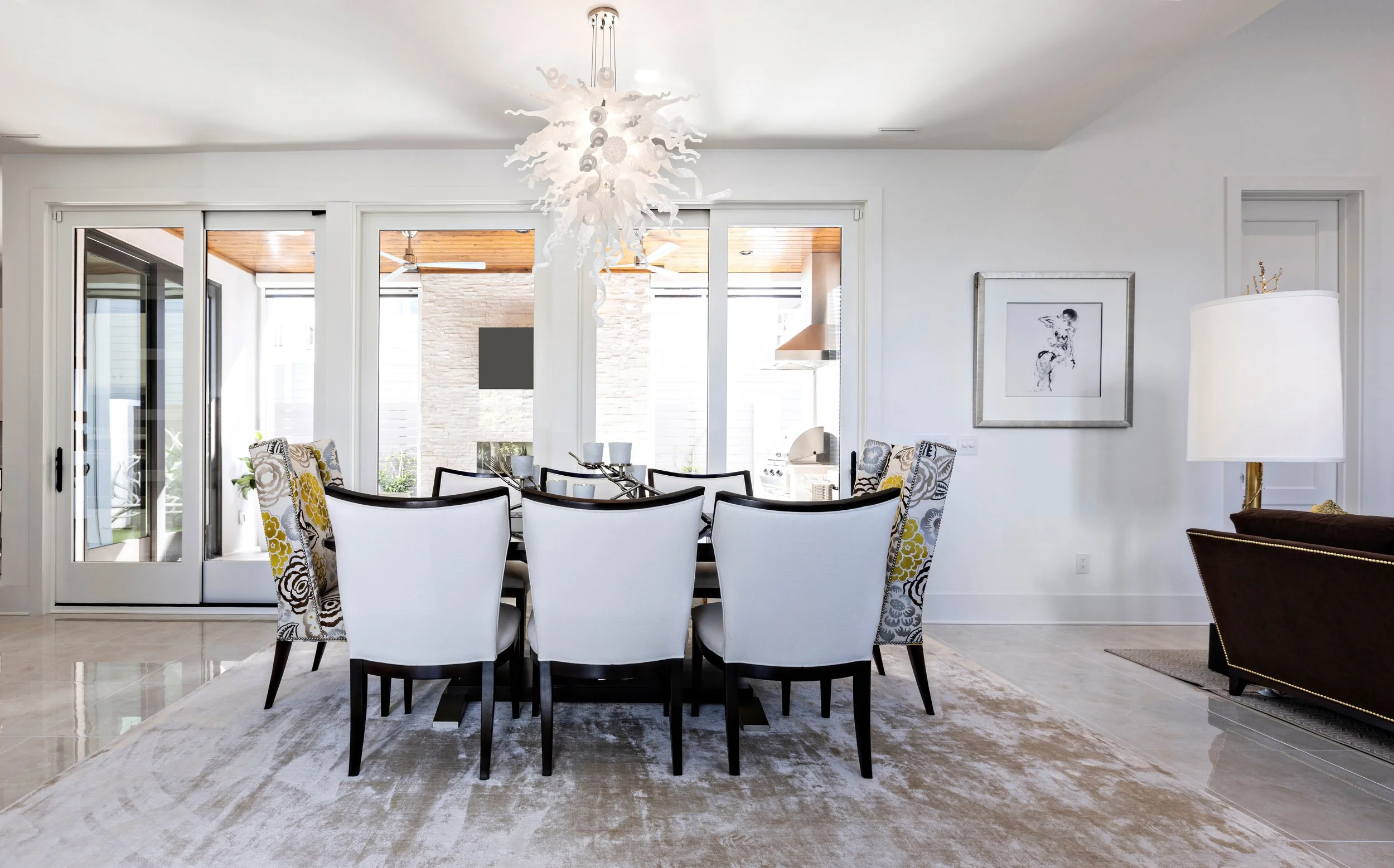 Modern dining room with a black table, white and patterned chairs, a chandelier, sliding glass doors leading to an outdoor patio, a framed art piece on the wall, and a seating area with a lamp.