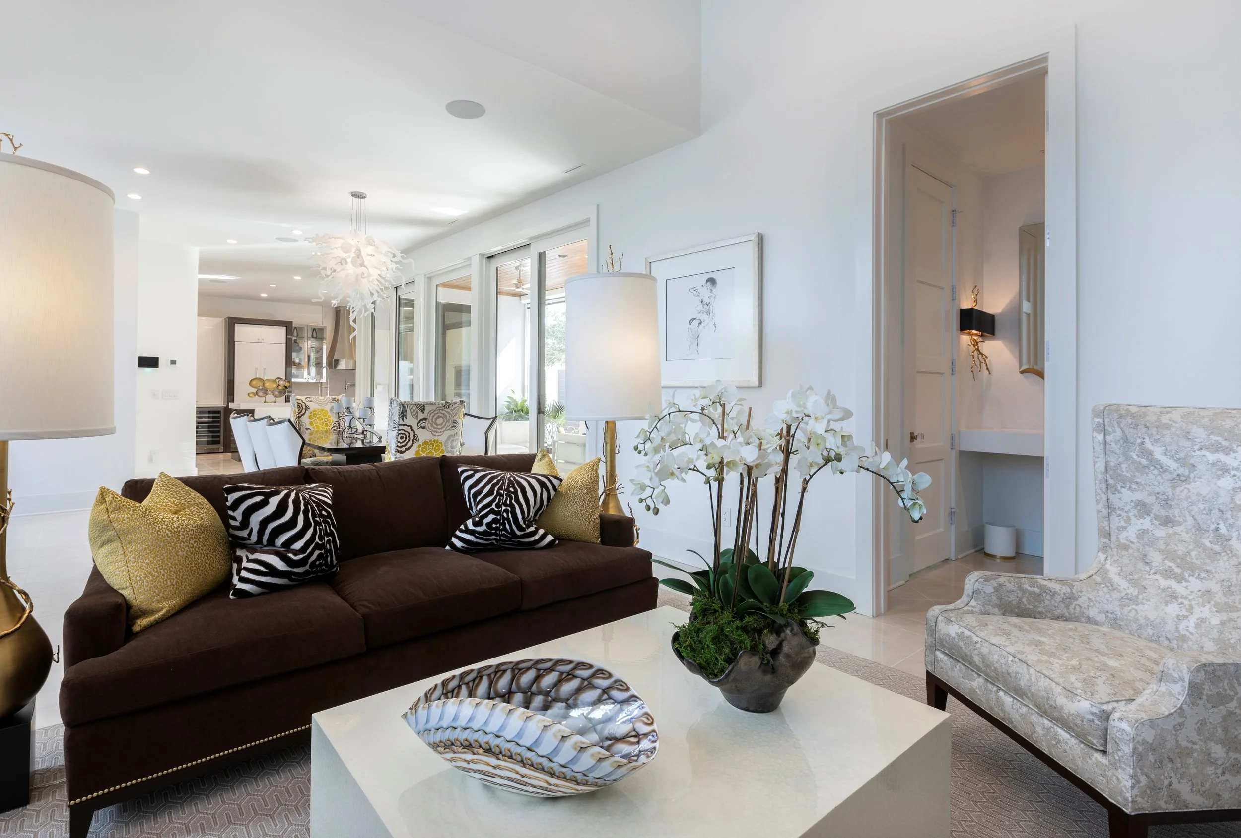 Living room with dark brown sofa decorated with gold and zebra-patterned pillows, large white orchid plant on a white coffee table, off-white armchair, white walls, contemporary art, and sliding glass doors leading to an outdoor patio.