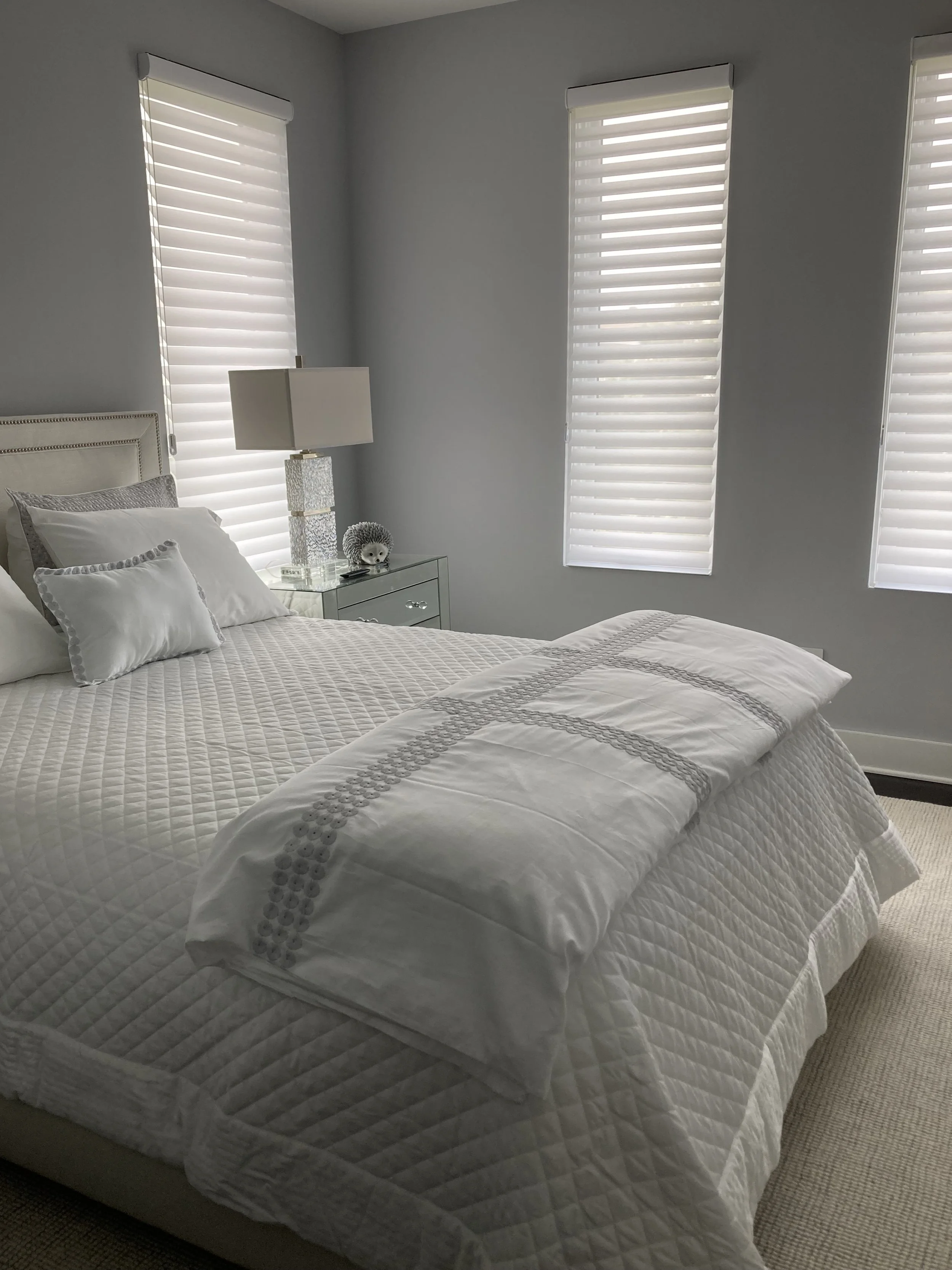 A cozy bedroom with light gray walls, three large windows with white horizontal blinds, a bed with white bedding and decorative pillows, a glass nightstand with a table lamp, and a plush hedgehog toy.