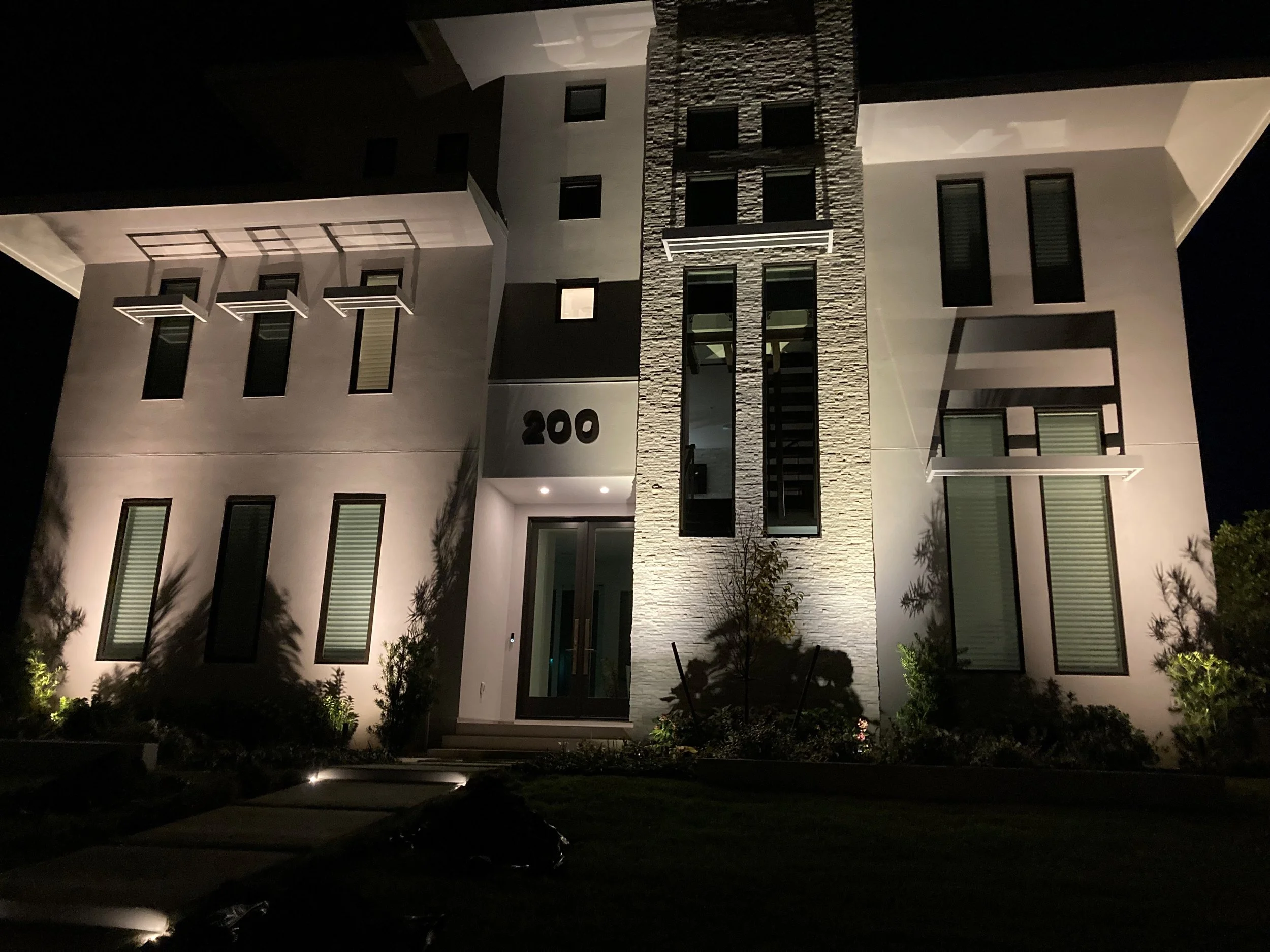 Night view of a modern multi-story residential building with illuminated exterior lighting, featuring large windows, balconies, and a stone accent wall near the entrance with house number 200.