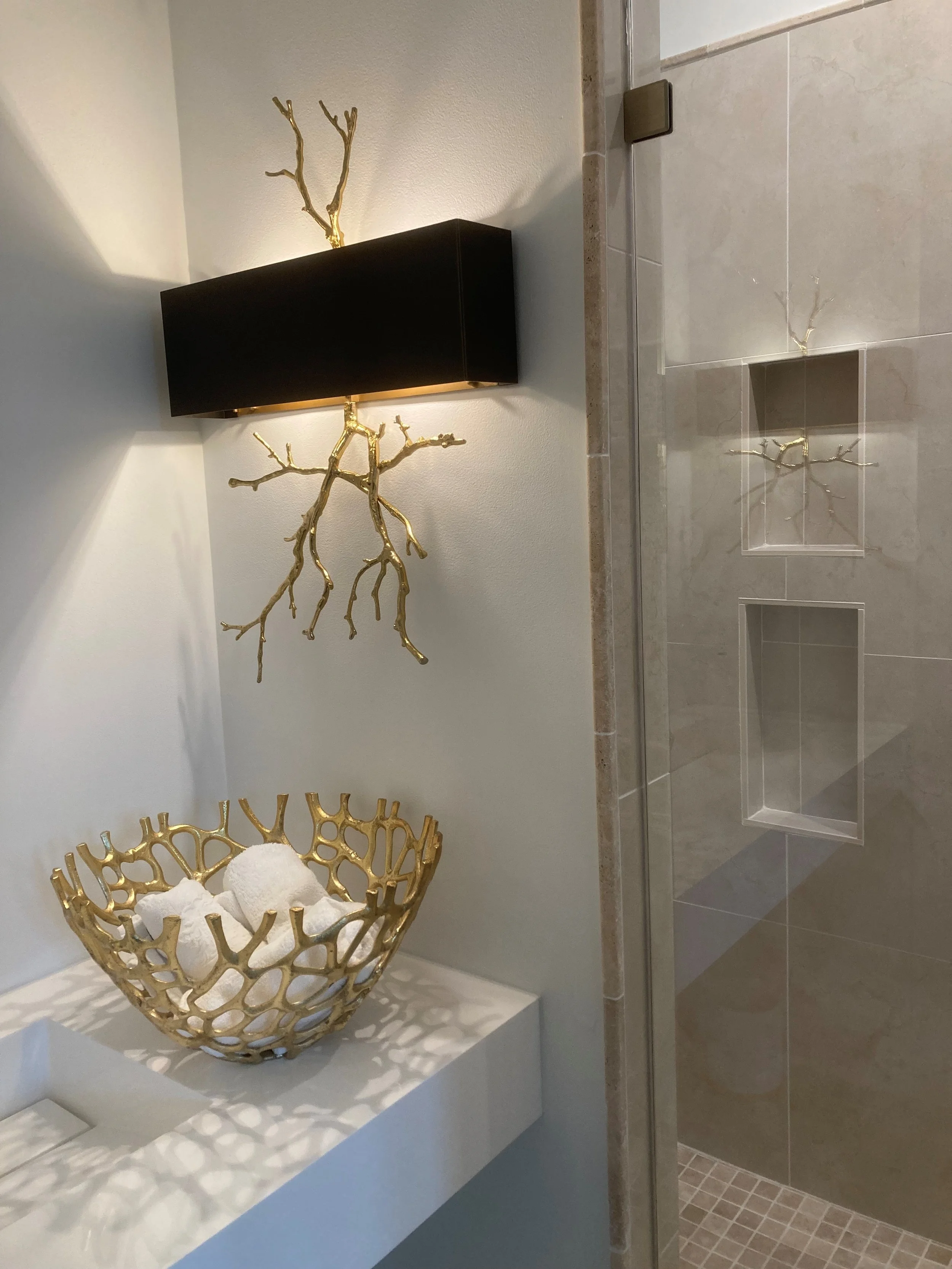 Decorative bathroom shelf with a gold abstract branch sculpture above a gold openwork fruit basket holding white bath bombs, and a shower with built-in niches, all with neutral tile and wall background.