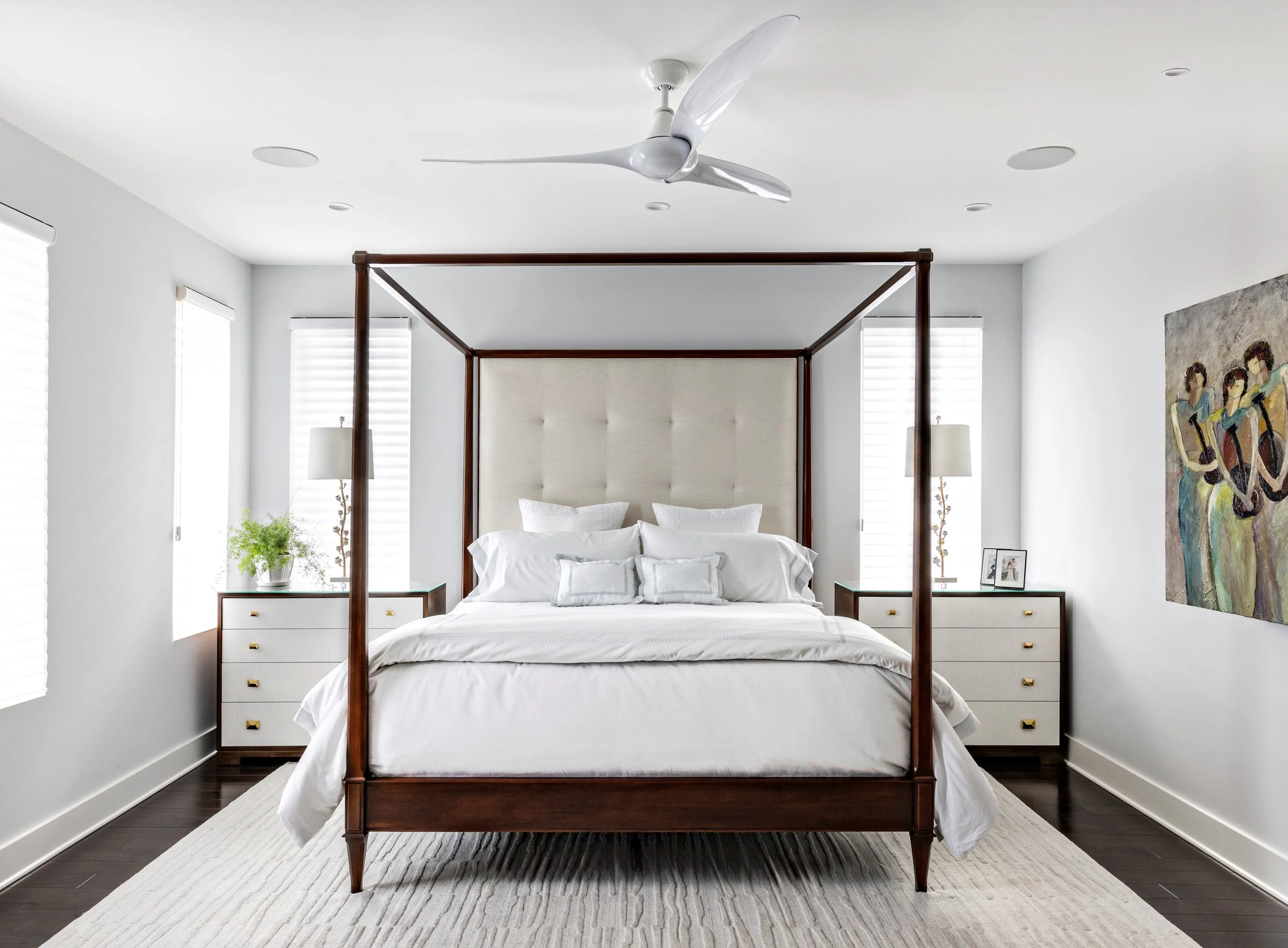 A modern bedroom featuring a four-poster wooden bed with a cream upholstered headboard, white bedding and pillows, two white nightstands with lamps and framed photos, a colorful wall art on the right, and a ceiling fan.