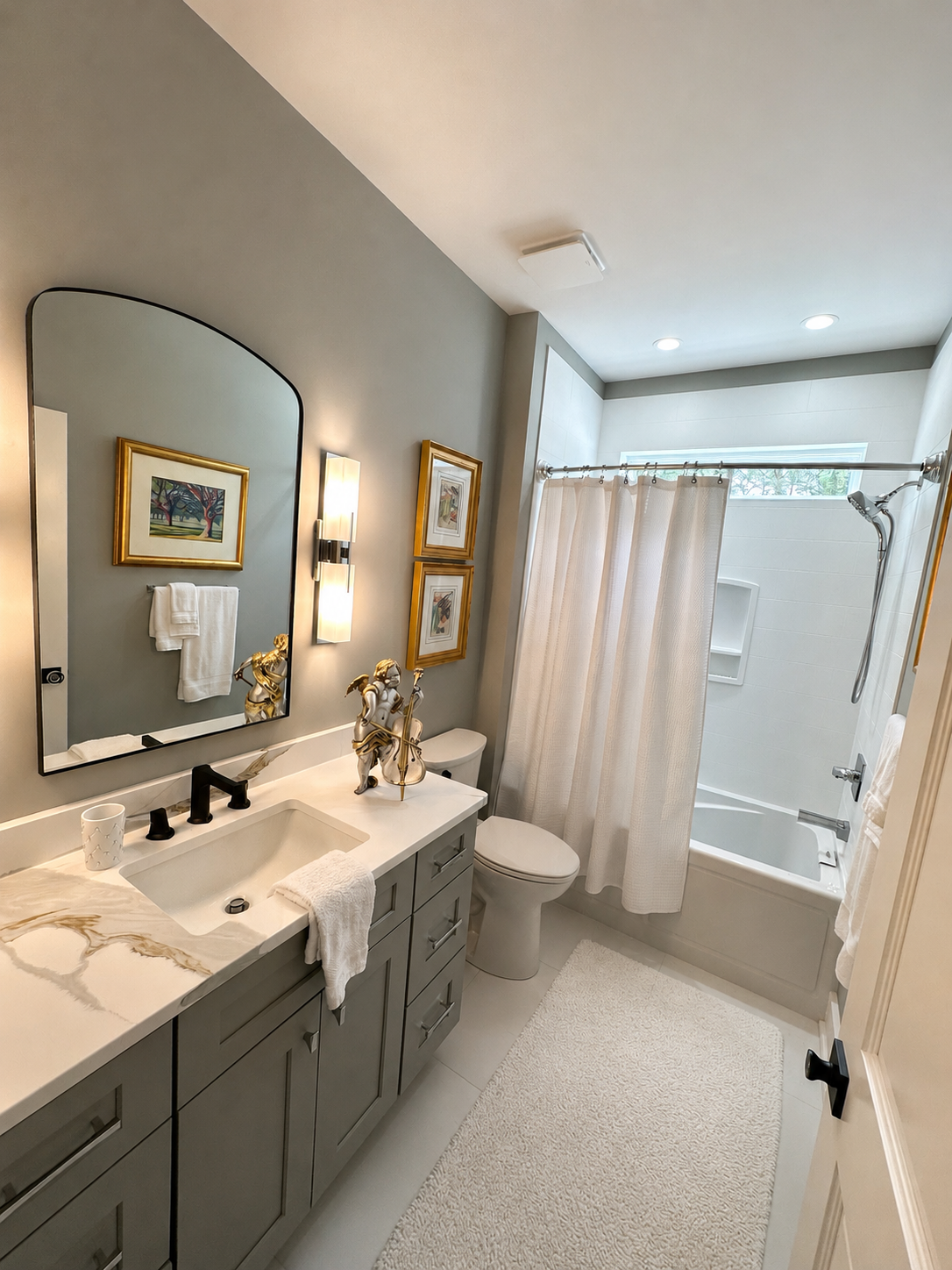A modern bathroom featuring a vanity with a marble countertop, a large mirror, and gold framed artwork; a white toilet; a bathtub with a shower, a cream shower curtain, and a small window; beige rug on the floor, and contemporary wall lighting.
