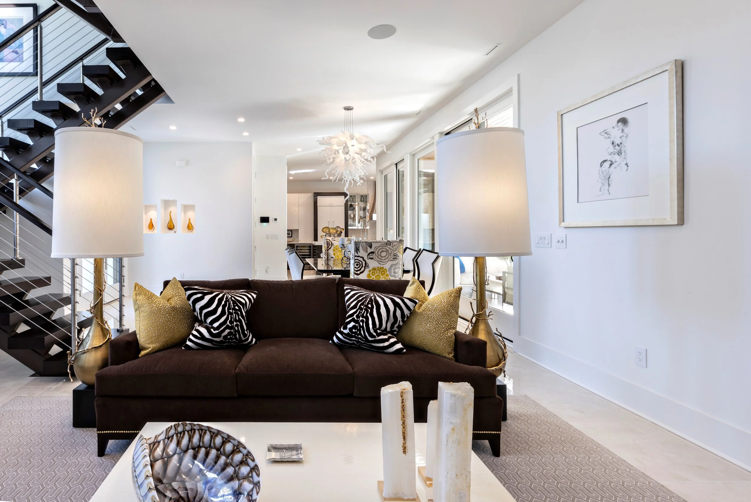 Modern living room with a dark brown sofa, zebra-patterned and gold decorative pillows, white walls, large windows, and various contemporary decor items.