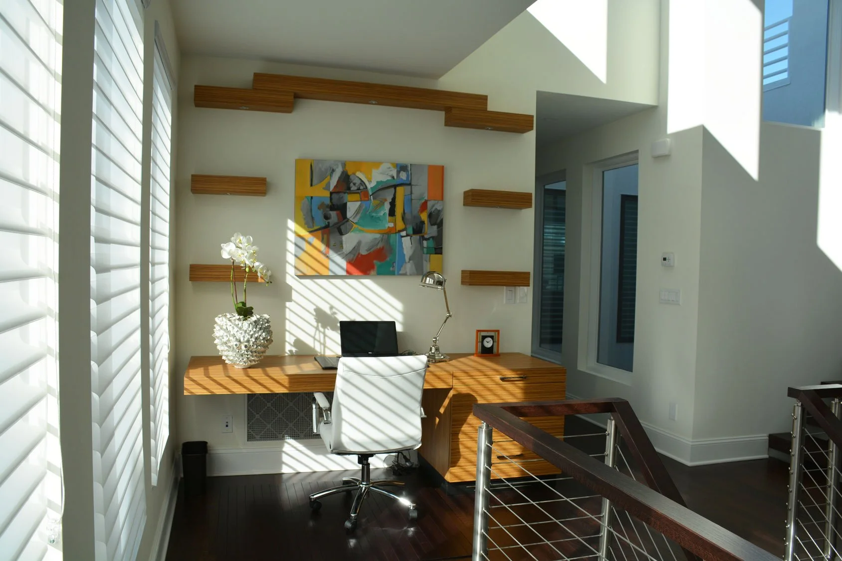 Modern home office with a wooden desk, white office chair, abstract art on the wall, and sunlight through window blinds casting shadows.