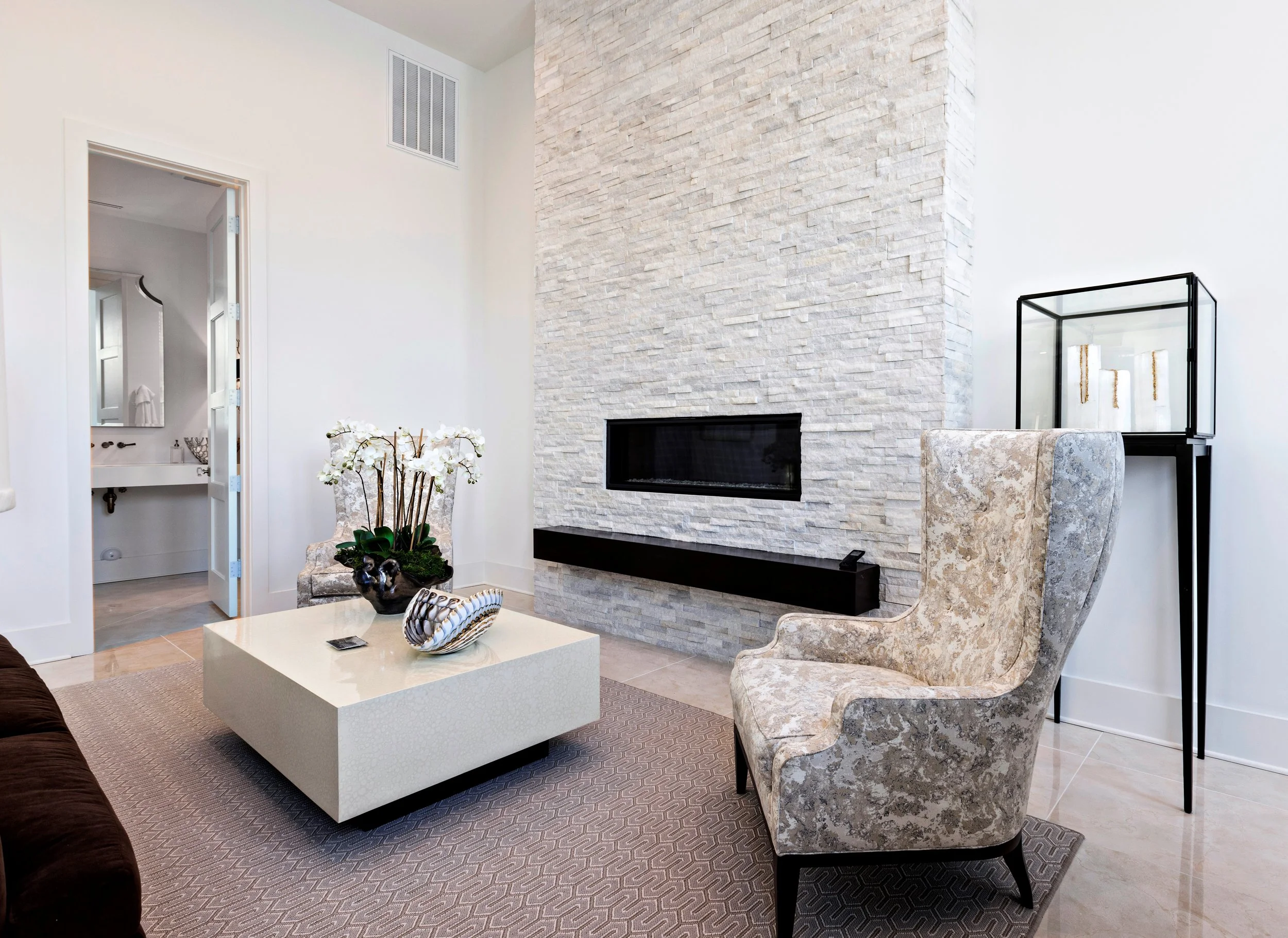 Living room with white textured stone fireplace, beige patterned armchair, black-framed glass display case, and white coffee table with decorative items.