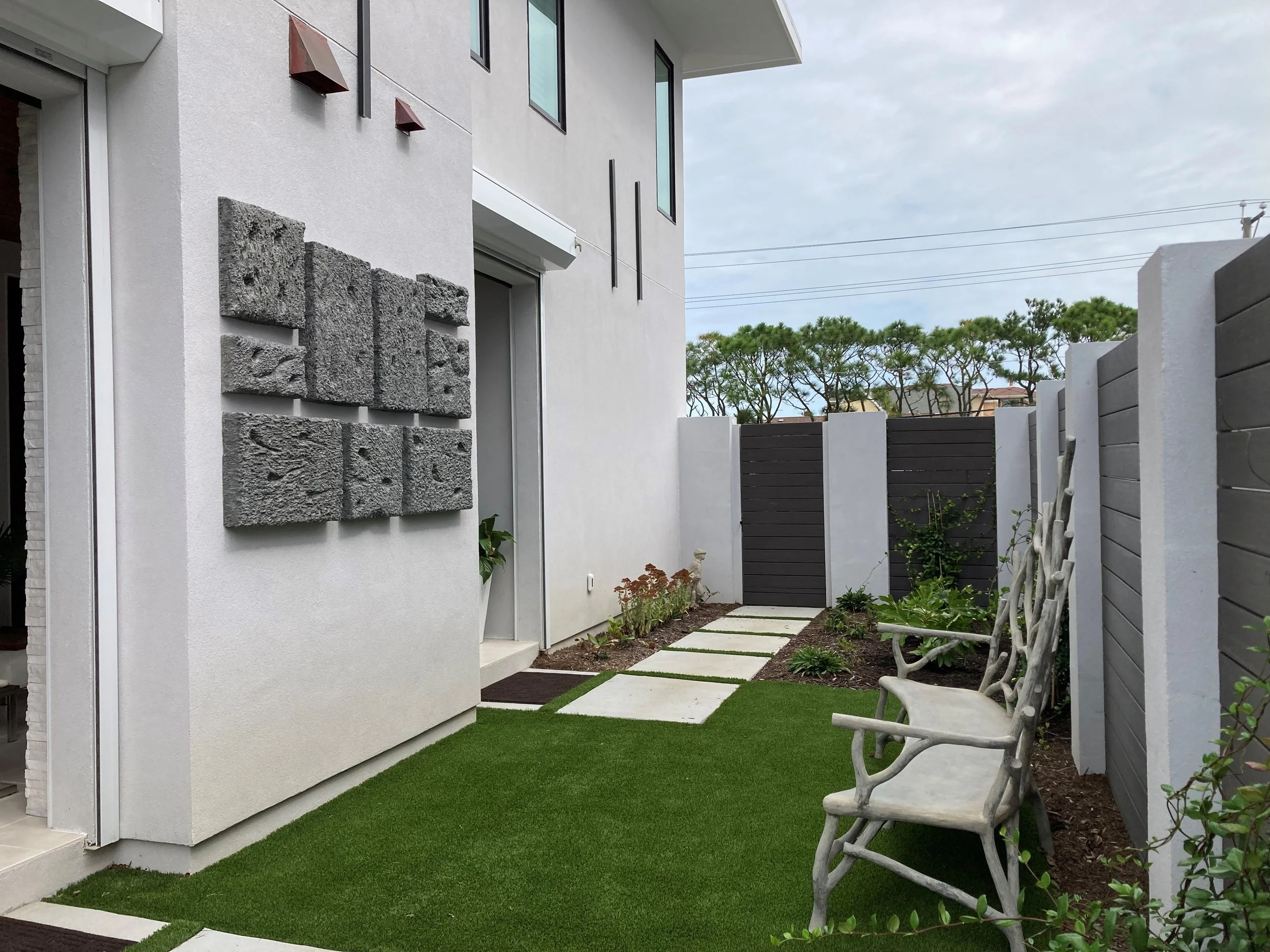 Modern backyard with white walls, decorative gray stone panels, a green artificial turf lawn, stepping stones path, garden plants, and a white bench with a tree branch design.