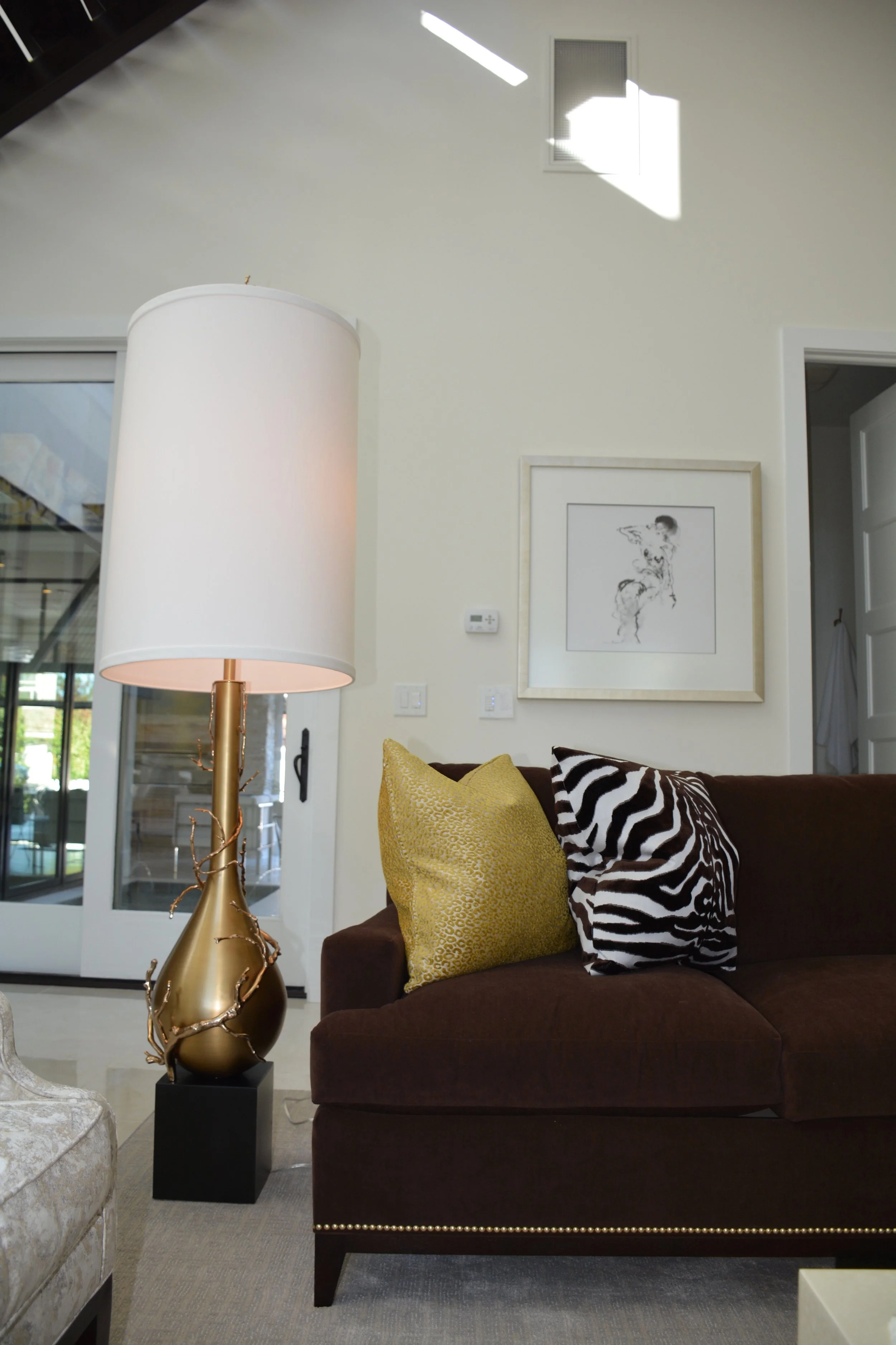 Living room with a dark brown couch, gold and zebra print pillows, a tall white floor lamp with a gold base, and a framed black-and-white sketch on a wall.