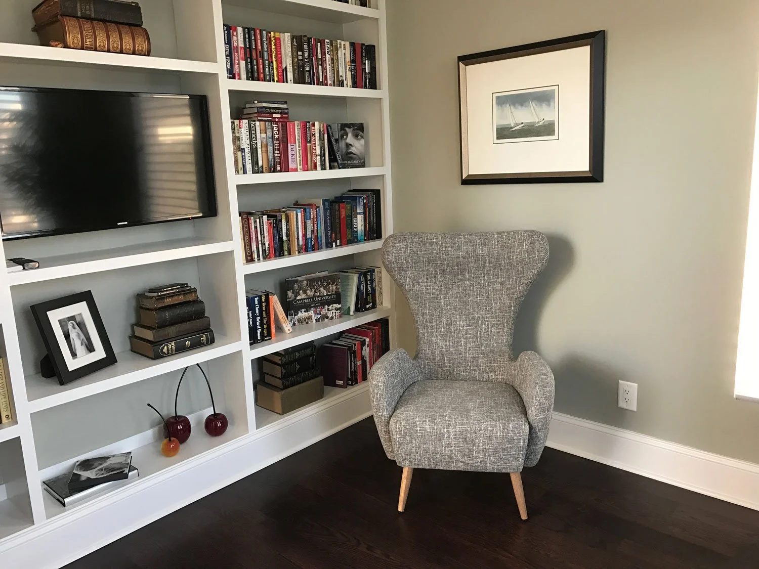 A cozy reading corner with a tall, wingback gray armchair next to a white bookshelf filled with books and decorative items, a framed picture of sailboats on a wall, and a television mounted on the bookshelf.