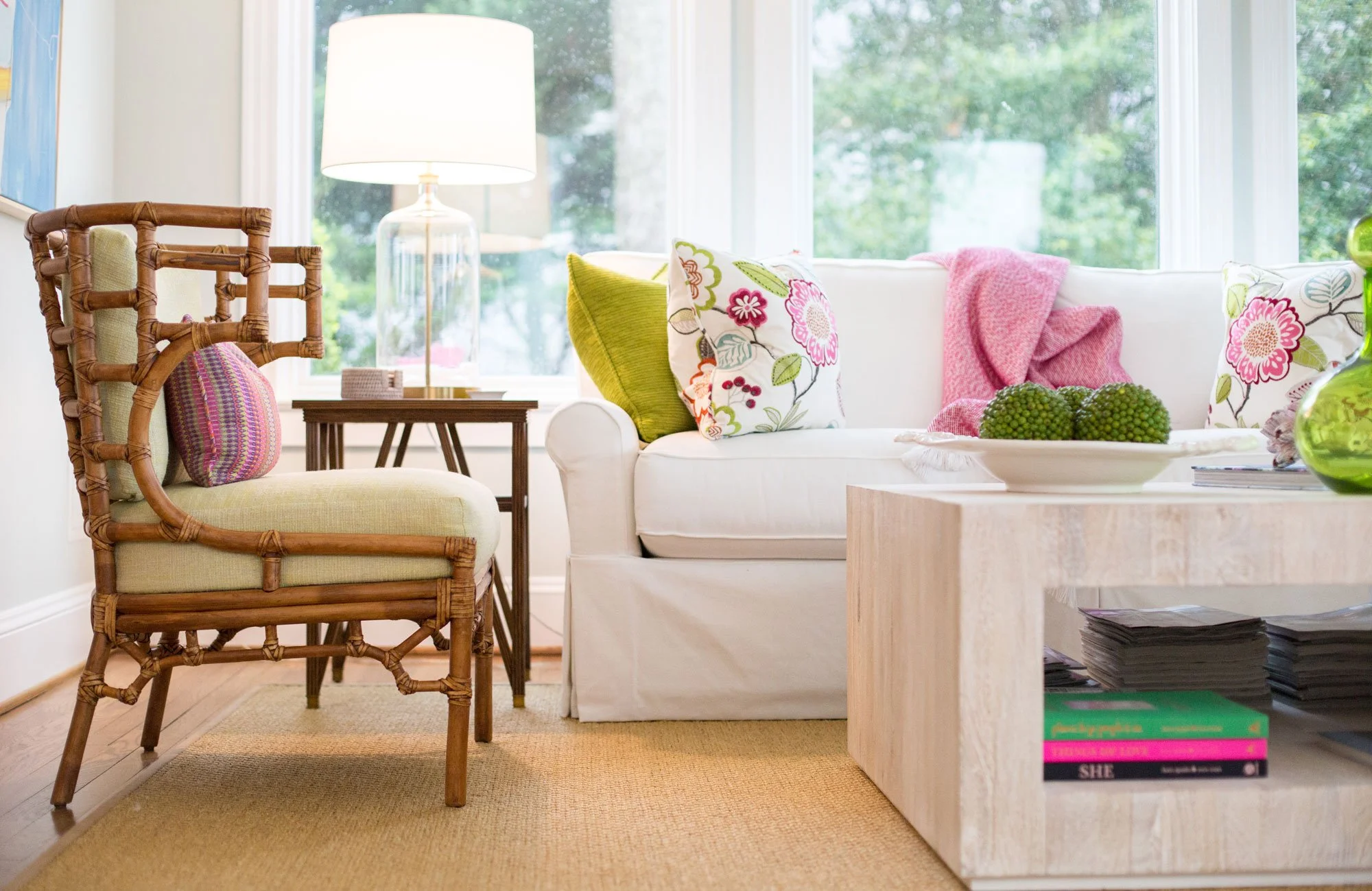 A living room area with a white sofa, colorful floral pillows, a pink throw blanket, a rattan armchair with a pink cushion, a wooden side table with a lamp, a large window with greenery outside, a beige rug, and a wooden coffee table with books and a