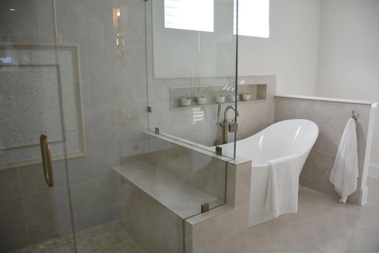 Bathroom with a glass shower enclosure, a bathtub, and beige tiled walls and floor. There are four small bowls on a built-in shelf, a towel hanging on a stand, and a white towel draped over the bathtub.