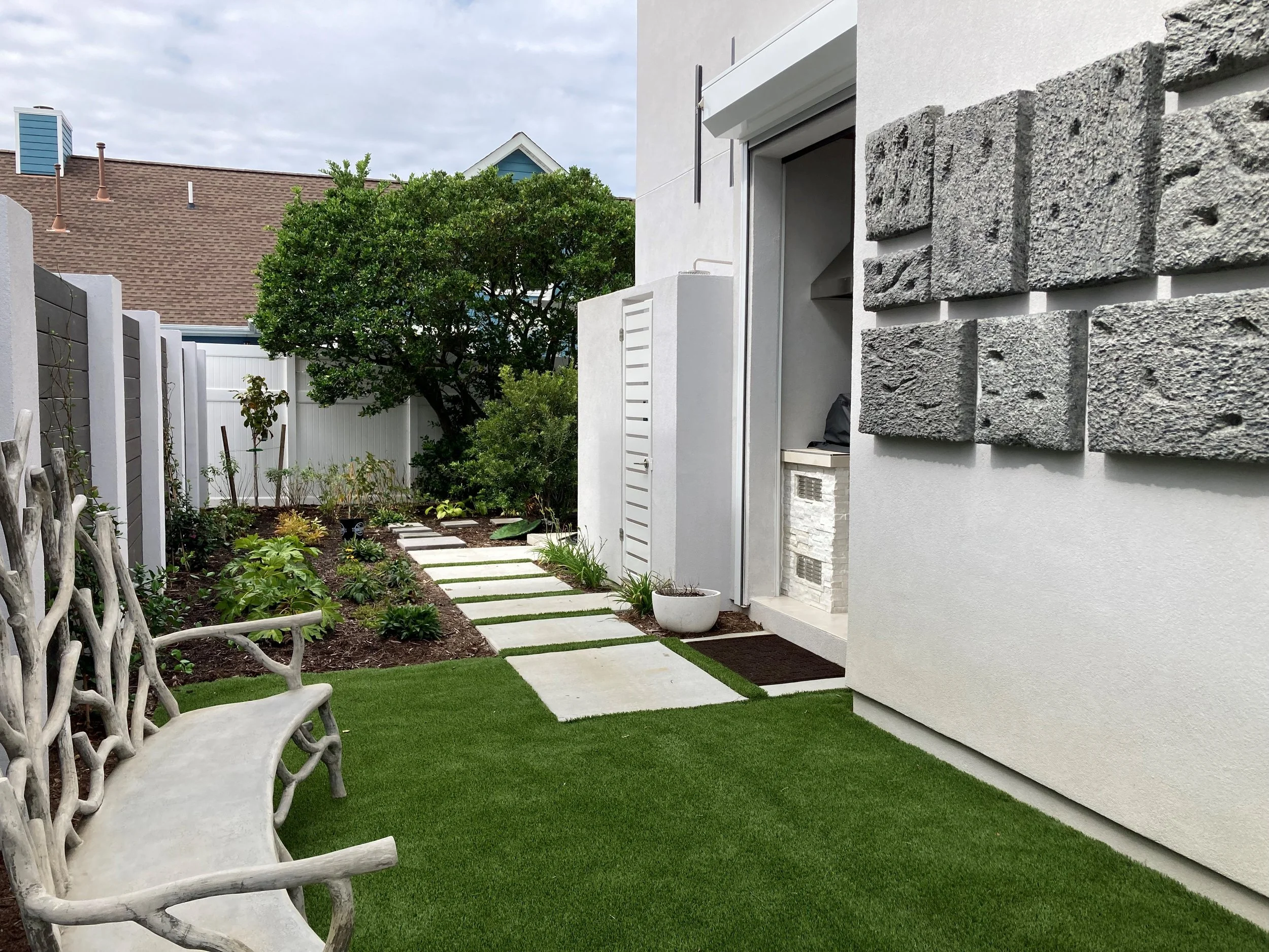 A small backyard garden with a stone pathway, green grass, a tree, and plants, surrounded by white fences and modern houses.