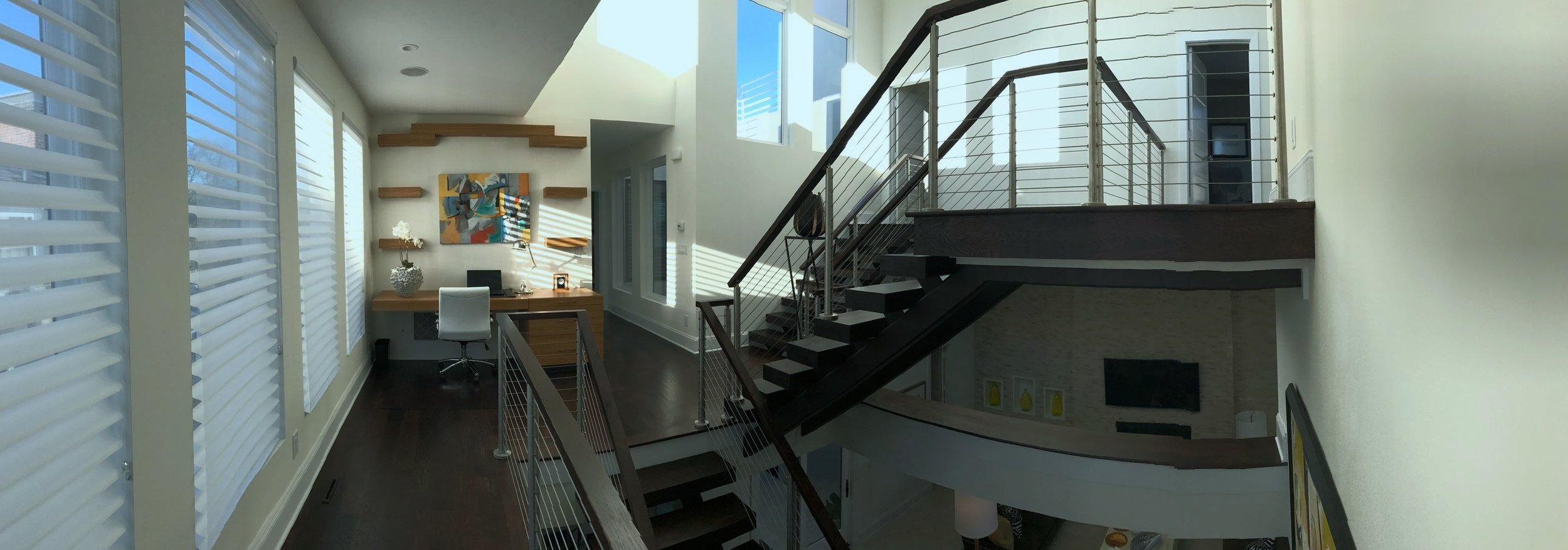 Modern interior of a two-story home with a staircase, a desk with a chair, large windows with blinds, and artwork on the wall