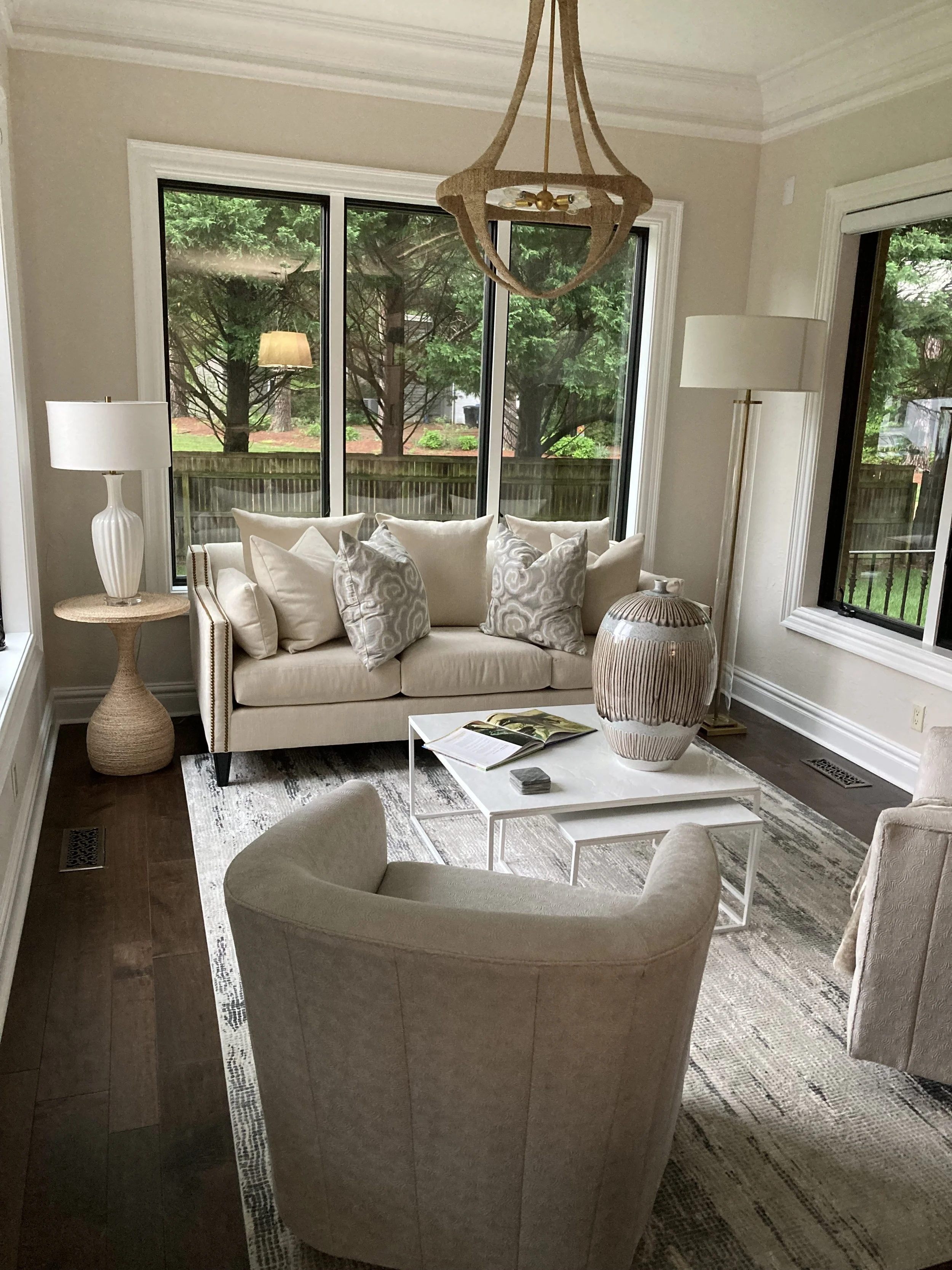 Living room with beige sofa, armchair, large window, wooden floor, round side table with lamp, white coffee table, decorative vase, and rug.