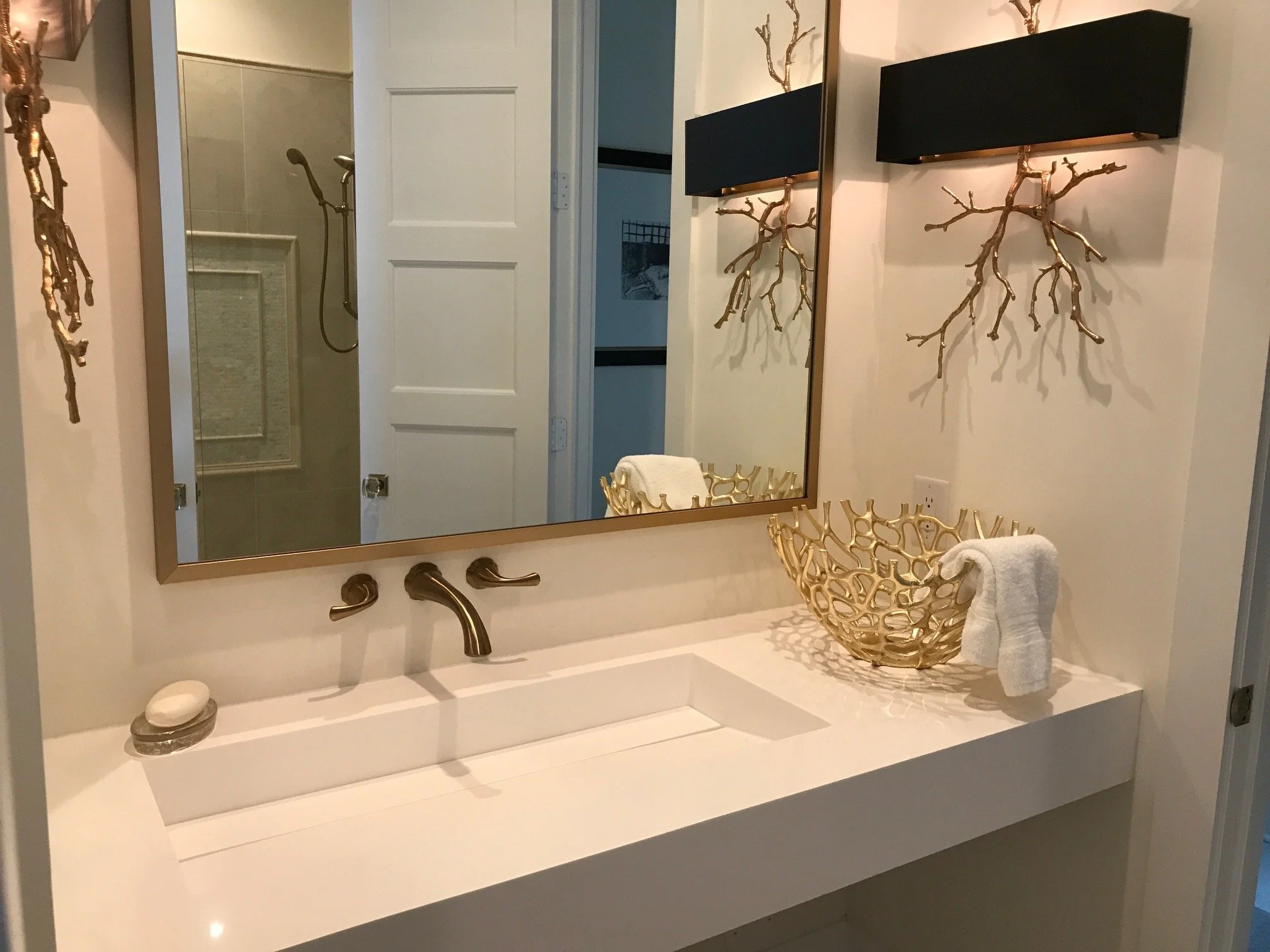 Bathroom vanity with a large mirror, gold branch-shaped wall decor, a gold decorative bowl with a white towel, and a soap dish, with a glimpse of a shower in the reflection.