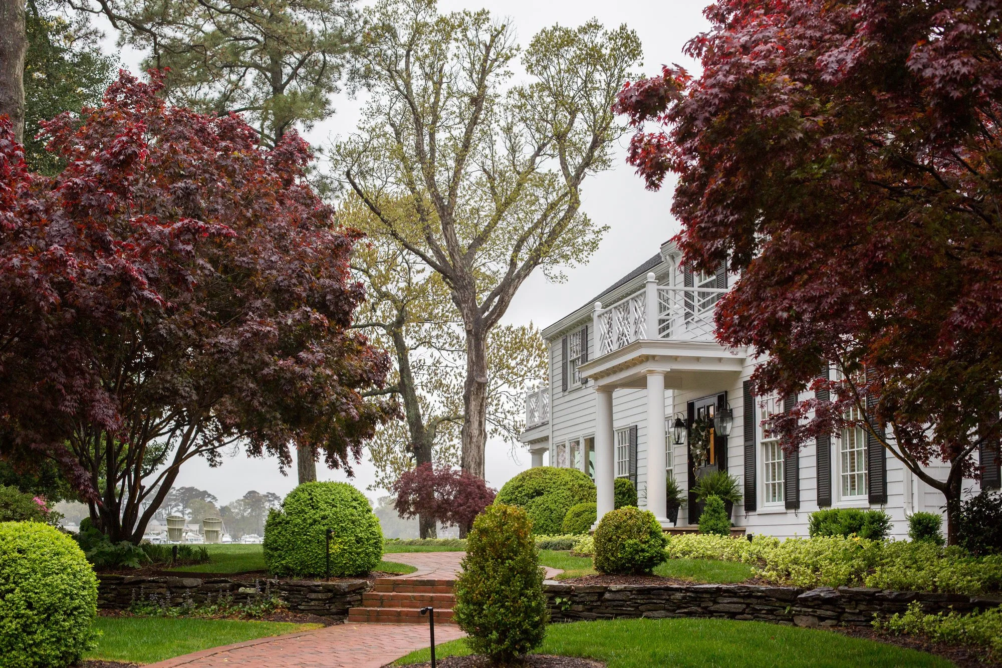 A white two-story house with black shutters, a porch with white columns, and a balcony, surrounded by well-maintained bushes and trees with red and green leaves, on a cloudy day.