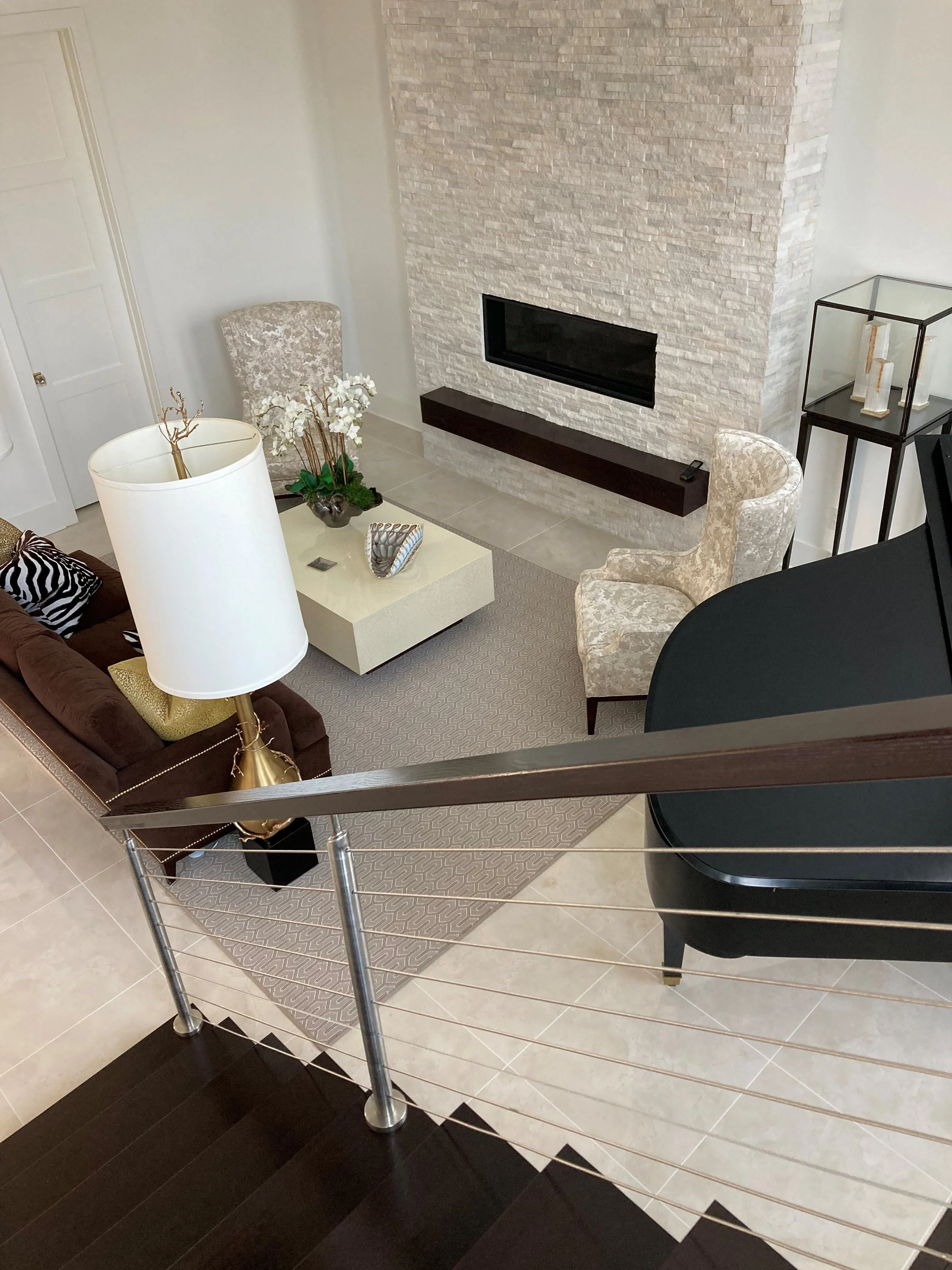 Top-down view of a modern living room with a fireplace, beige patterned armchairs, a white coffee table with decorative items, a plush brown sofa with pillows, a tall white lampshade, and a glass display case in the corner.
