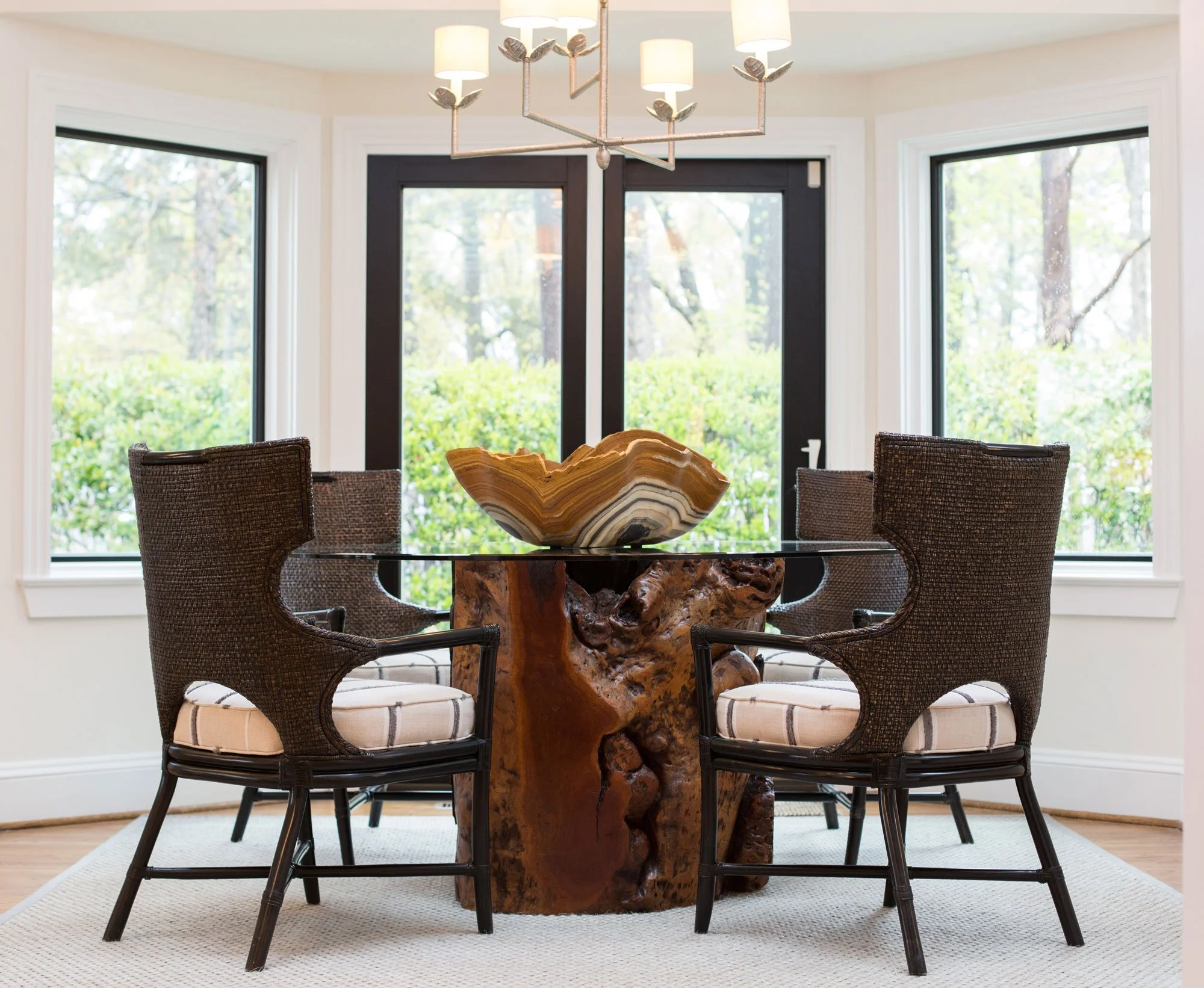 A round glass dining table with a unique wooden base, surrounded by four dark wicker chairs with light-colored cushions, in front of large windows with black frames and a view of green bushes and trees outside, with a modern chandelier hanging above.