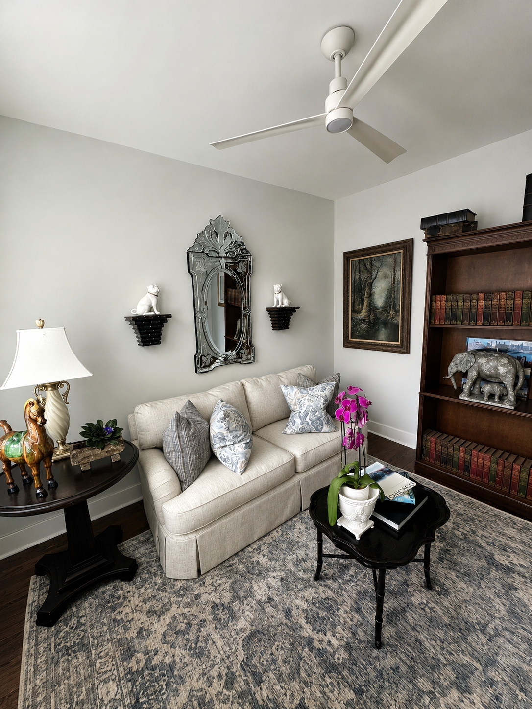 Living room with beige sofa, decorative pillows, black side table with horse figurine, lamp, orchid plant, dark wood bookshelf with animal statues, artwork, and framed mirror, ceiling fan, patterned area rug.