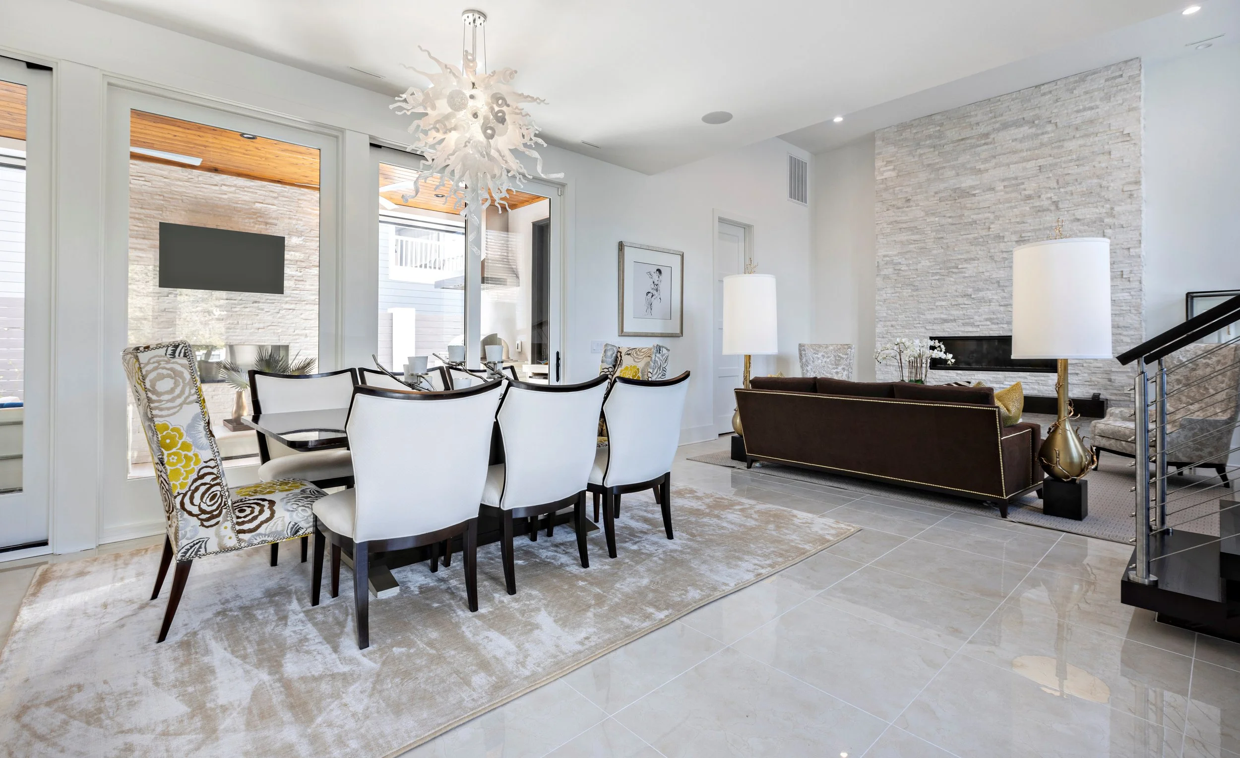 Open-concept living and dining area with white walls, tiled floor, dining table with white and patterned chairs, and a gray stone fireplace with a mounted TV above. Bright, modern decor with two large white lamps and artwork.