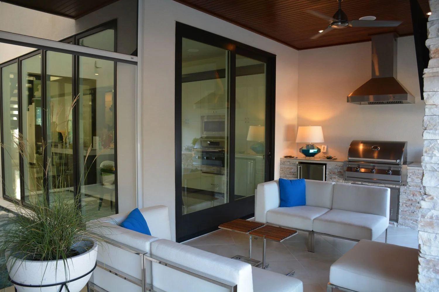 Outdoor patio with white sectional sofa with blue pillows, potted plant, and a built-in grill and counter with lamps, in a modern house with large glass windows and a wooden ceiling.