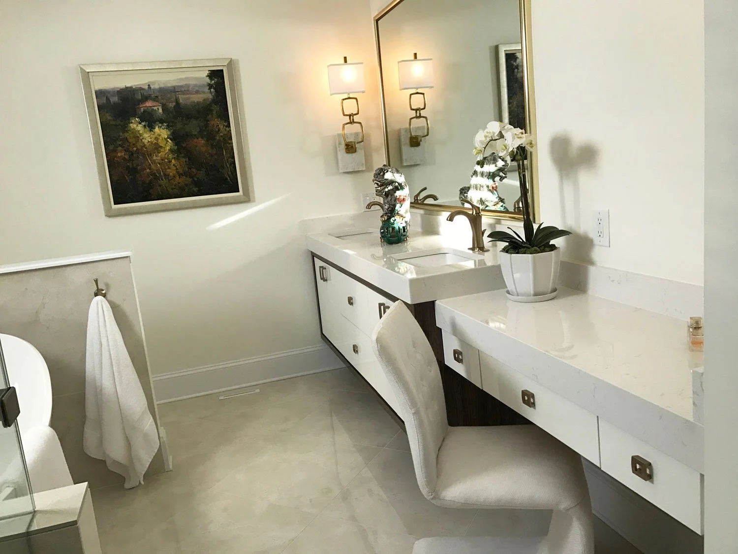 A modern bathroom with a white vanity featuring a marble countertop, a large mirror, and two decorative horse head sculptures. There is a white orchid in a pot on the counter, two wall-mounted lamps above the mirror, a painting of landscape on the wa
