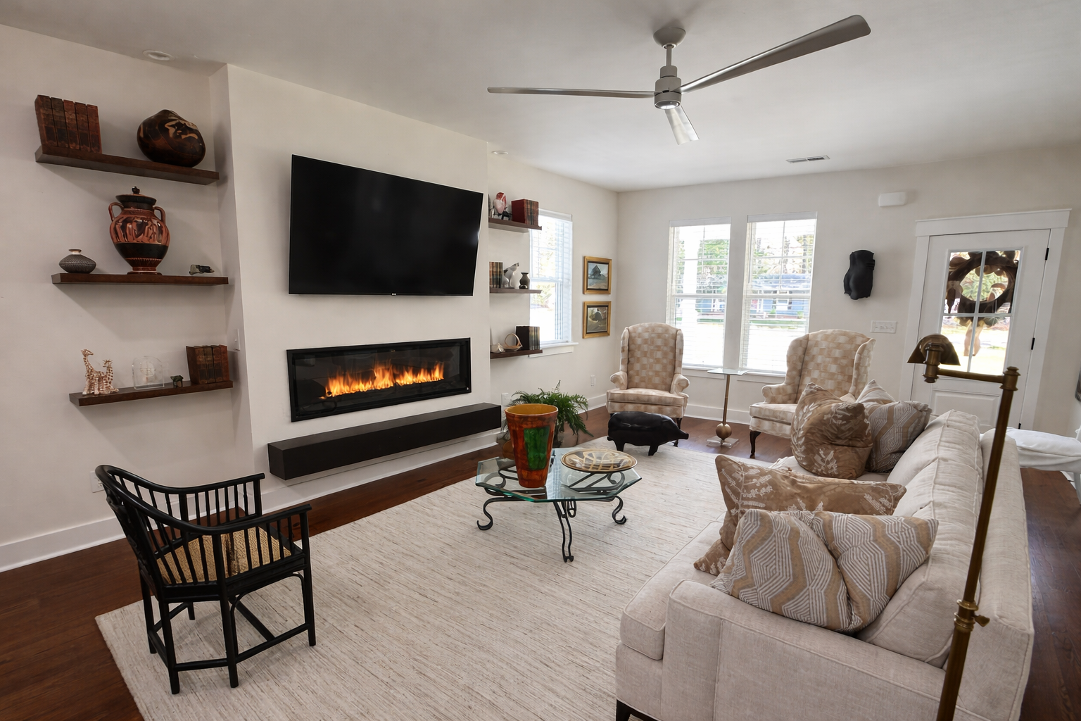 Bright living room with a light-colored sofa, two armchairs, a glass coffee table, a wall-mounted TV, a modern fireplace, wooden shelves with decorative objects, large windows, and a ceiling fan.