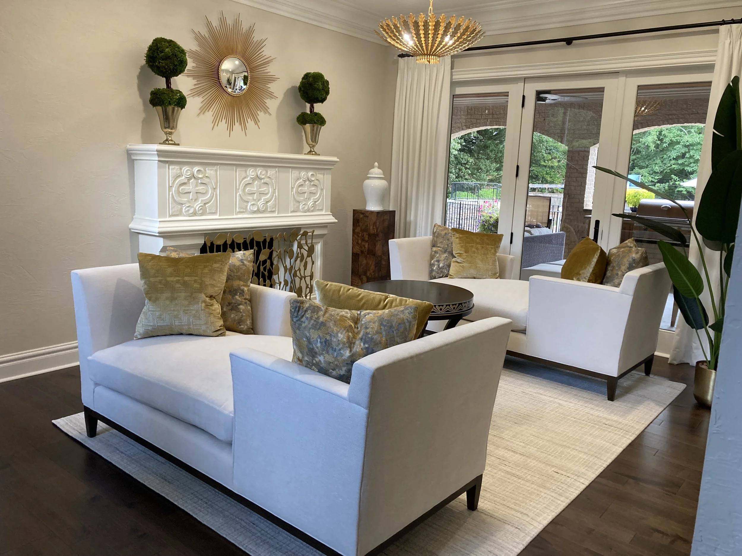 Living room with white sofas and gold pillows, dark wood table, fireplace with decorative white mantle, fireplace screen, and glass sliding door leading to outdoor patio with greenery.