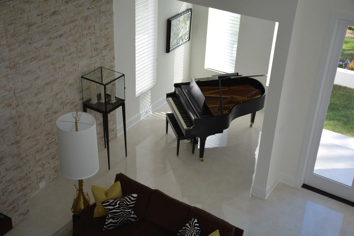 A living room with a black grand piano near a window, a brick accent wall, a white lampshade, two tall candle holders in a glass display case, a sofa with zebra-patterned pillows, and sunlight streaming through blinds.