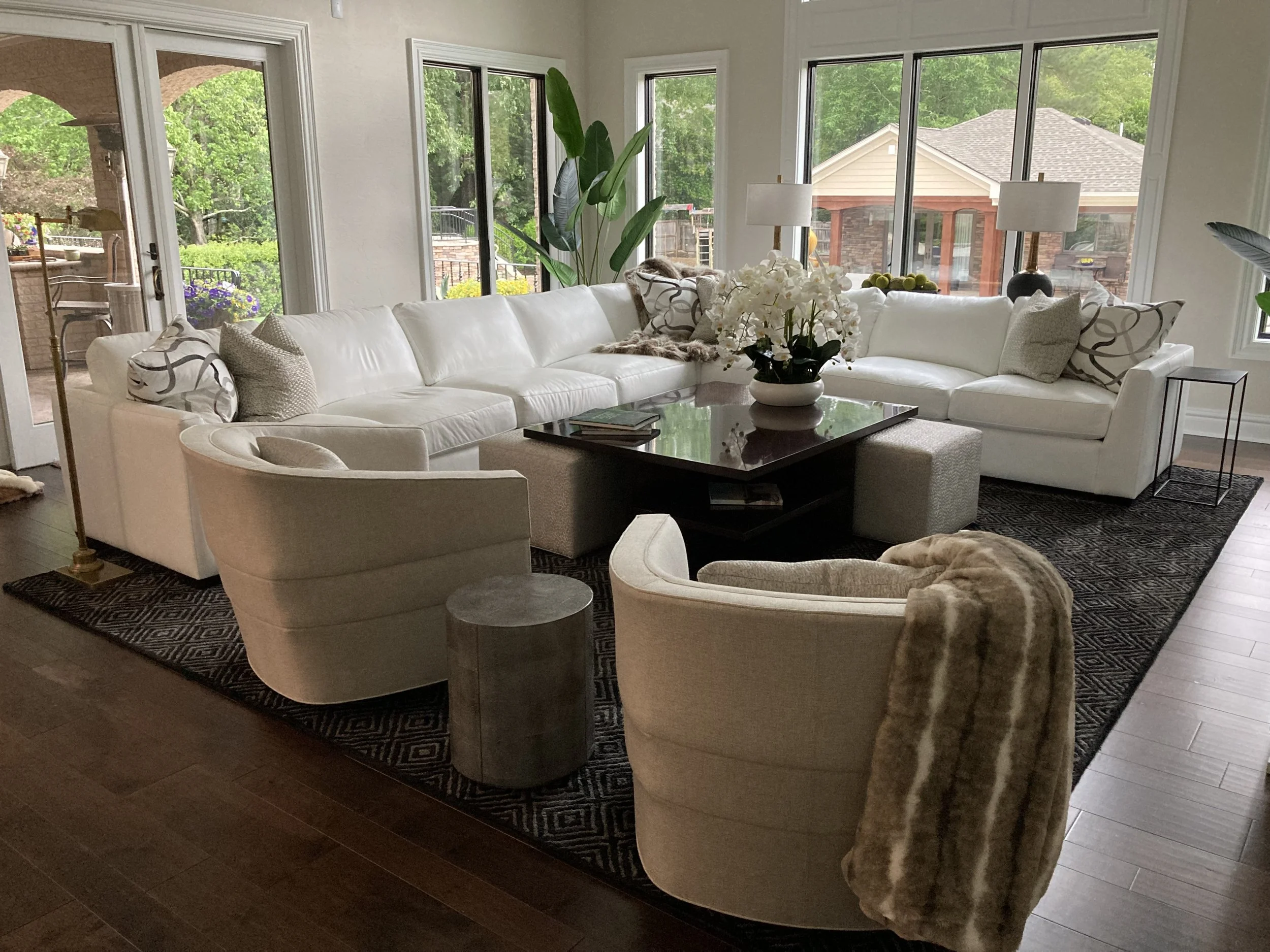 Living room with large white sectional sofa, two beige armchairs, a black coffee table, and a floral centerpiece. Large windows and sliding door reveal a garden with trees and a patio.