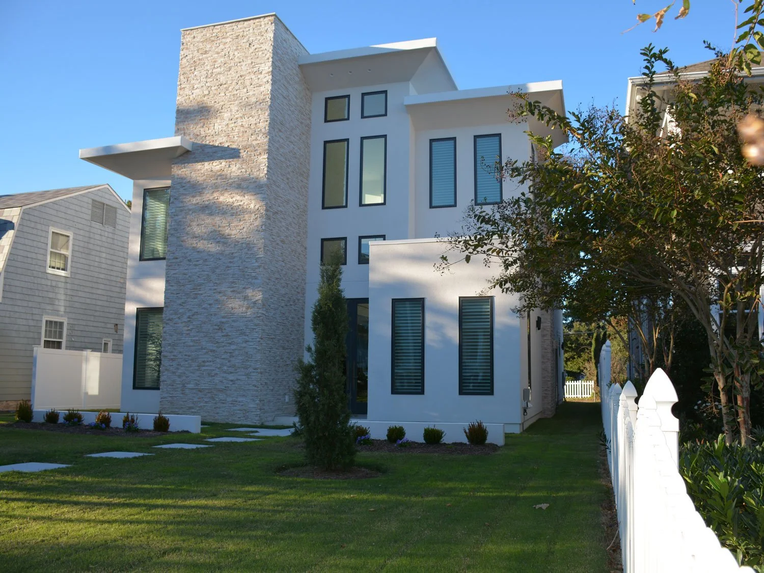 Modern white house with large windows and stone accents, surrounded by a green lawn, small bushes, and a white picket fence, under a clear blue sky.