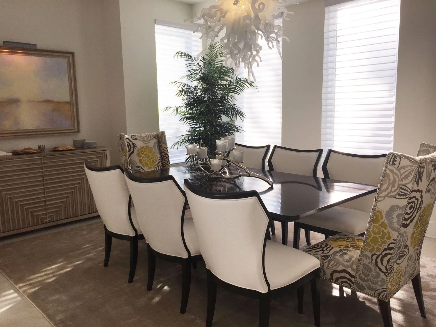 A dining room with an elegant black wooden table surrounded by eight white and patterned upholstered chairs. A large leafy green indoor plant is placed near the windows, which have white blinds. There is a decorative centerpiece with candles on the t