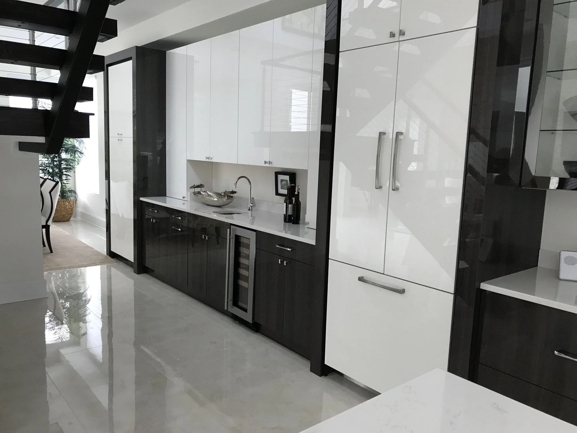 Modern kitchen with white and black cabinets, a small wine fridge, a sink, and counter space. Part of a dining area with a chair and plant visible.