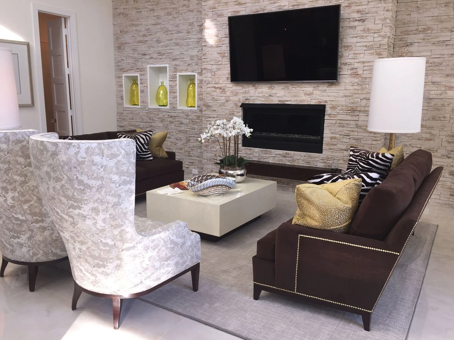 Living room with beige stone wall, brown and white sofas, patterned pillows, cream armchair, coffee table with decorative bowls and white flowers, large white floor lamp, wall-mounted TV, and a built-in fireplace.