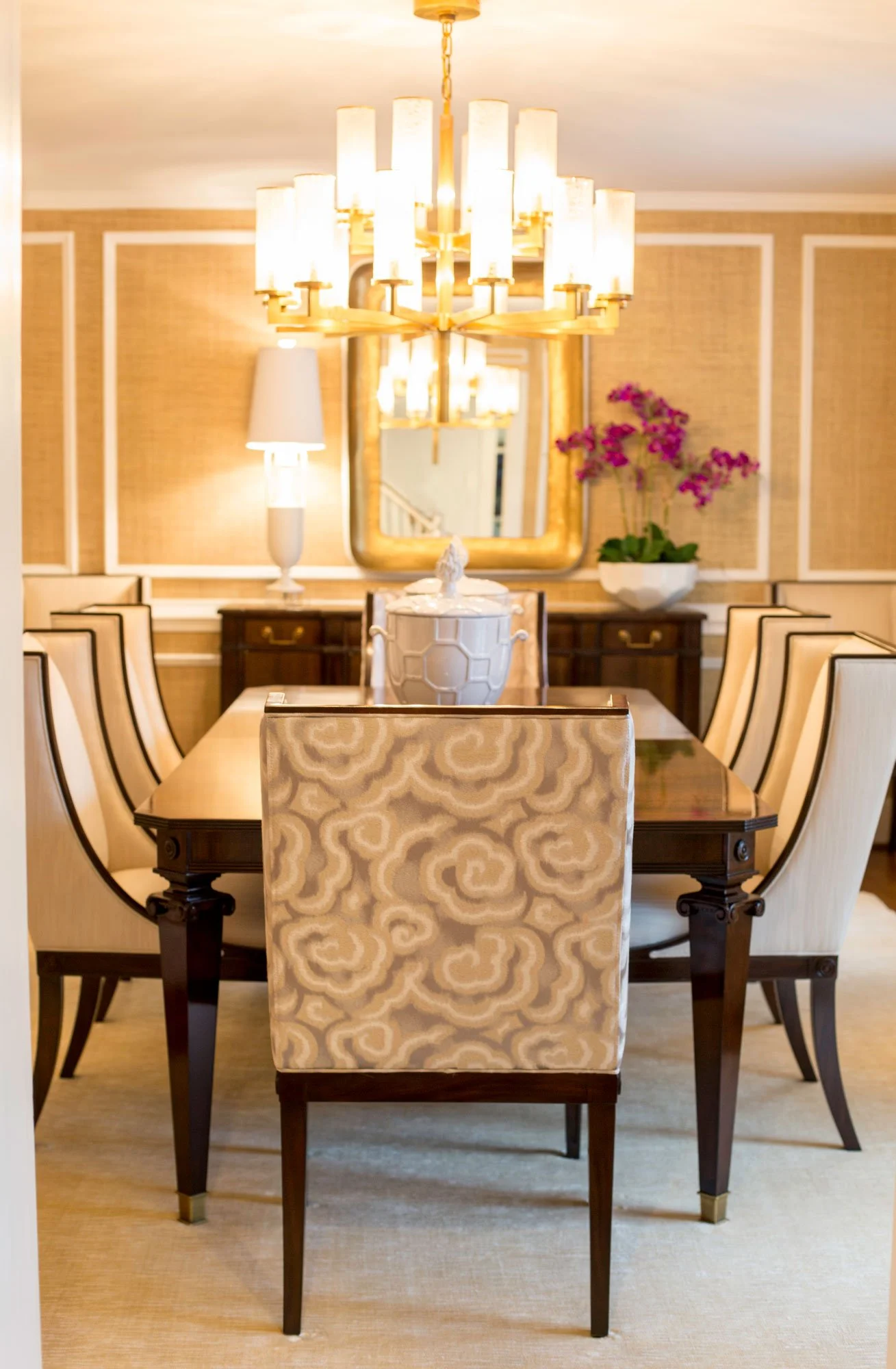 A formal dining room with a wooden table, cream-colored upholstered chairs, a large chandelier, a mirror, a lamp, a sideboard, and a vase of pink flowers.