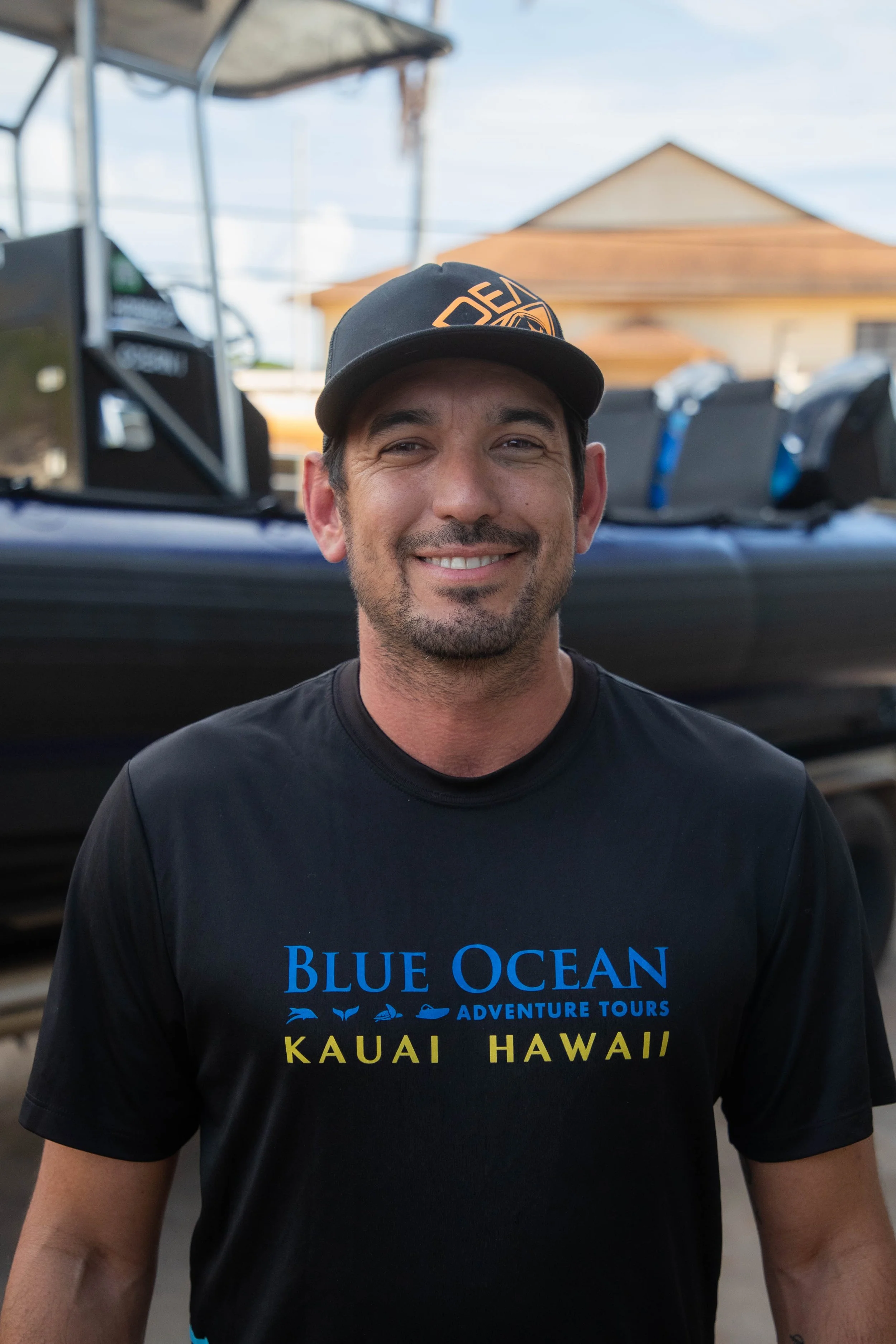 Smiling man in black "Blue Ocean Adventure Tours Kauai Hawaii" T-shirt and cap, standing outdoors with boat equipment and a house in the background.