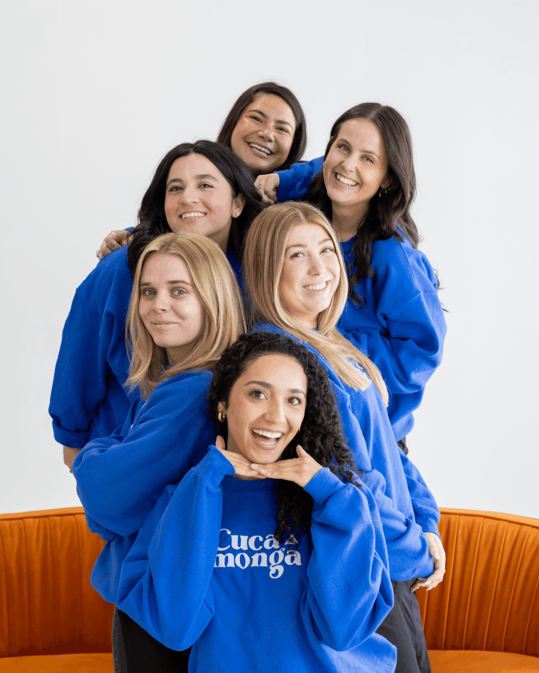Team photo of six women in matching blue Cucamonga Media sweatshirts, smiling and posed playfully on an orange couch.