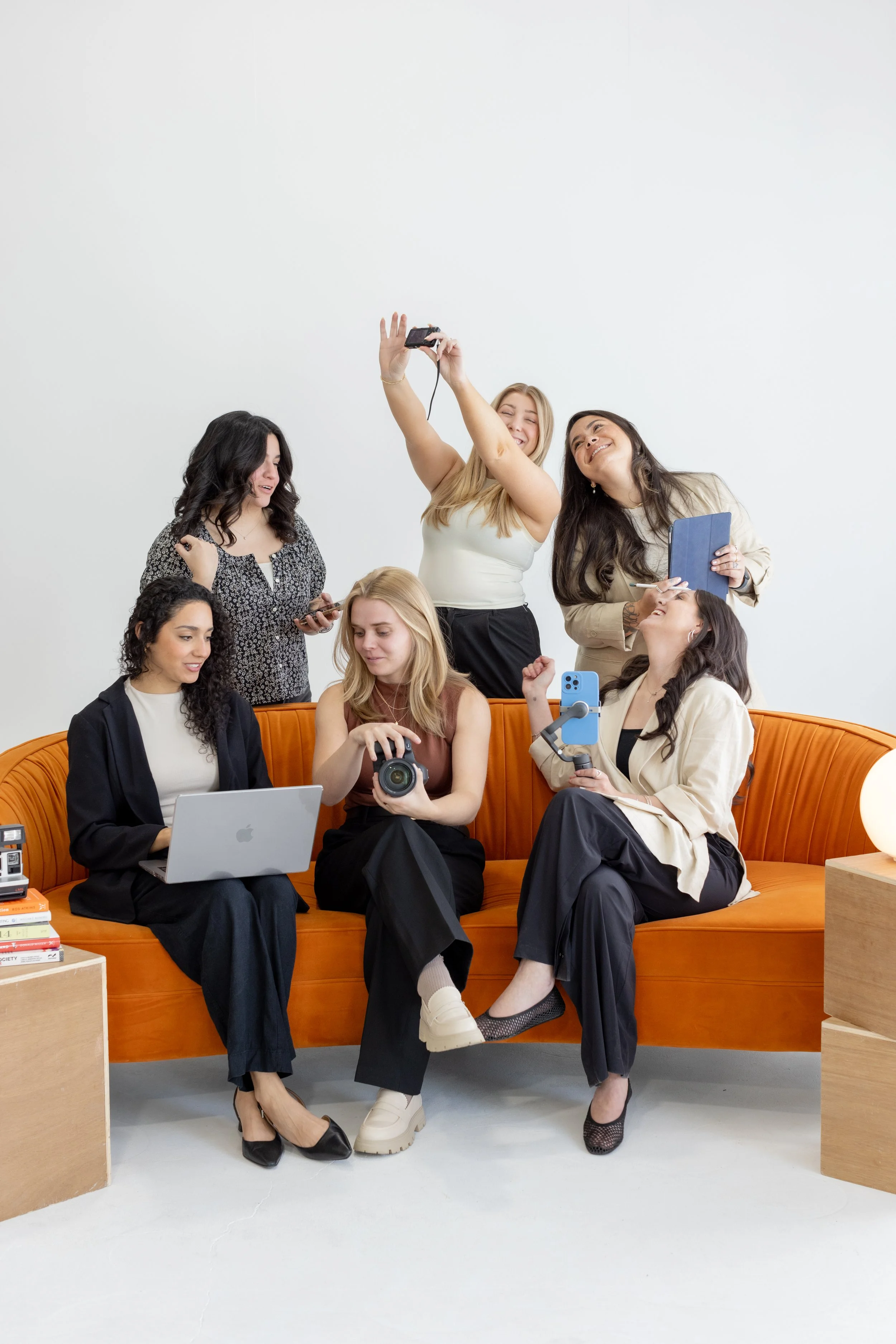 Cucamonga Media team members with a white background. Three team members are behind the couch taking a photo and three more are sitting on the couch conversing.