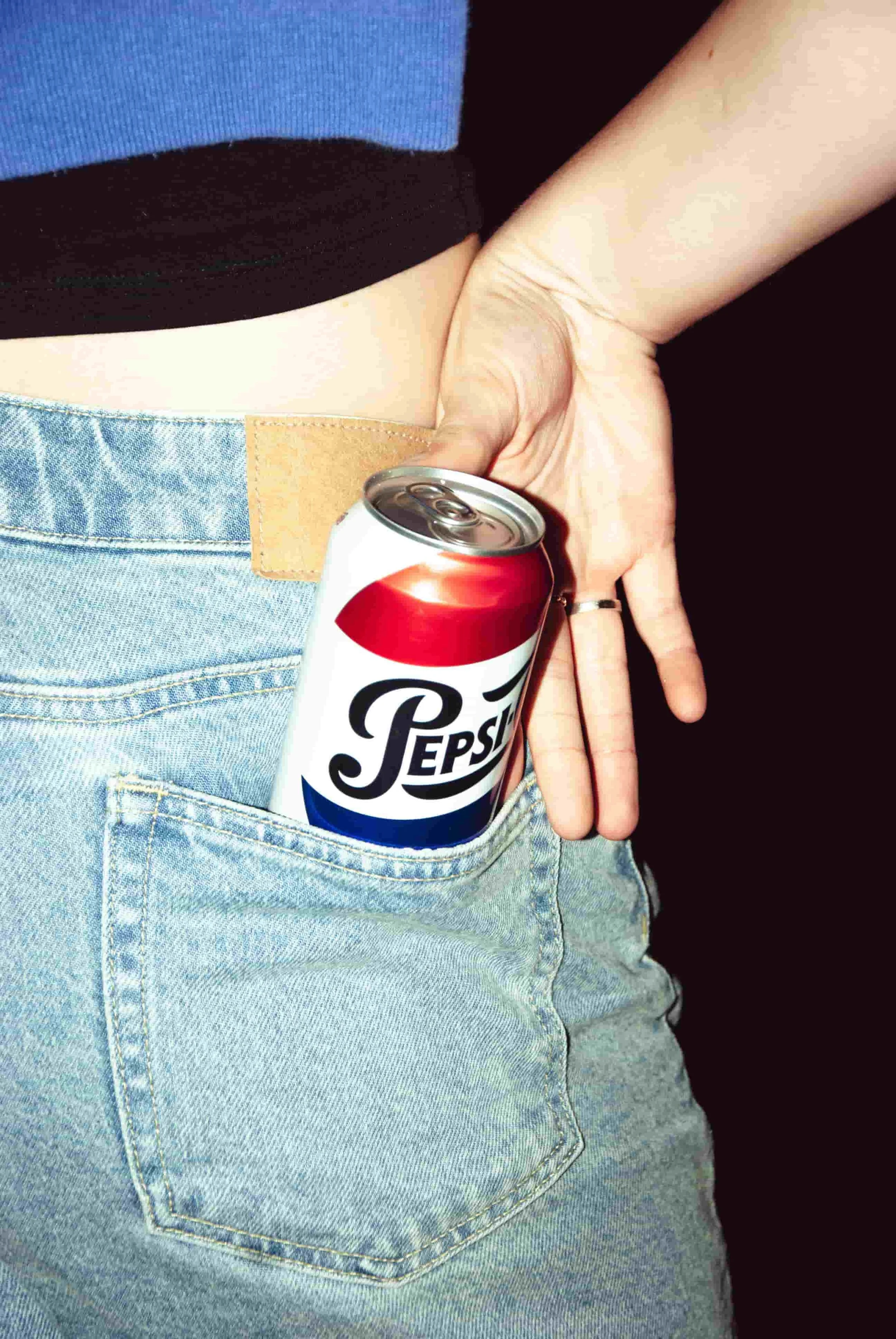 Close-up of a Pepsi can being pulled from the back pocket of light wash denim jeans, against a dark background.