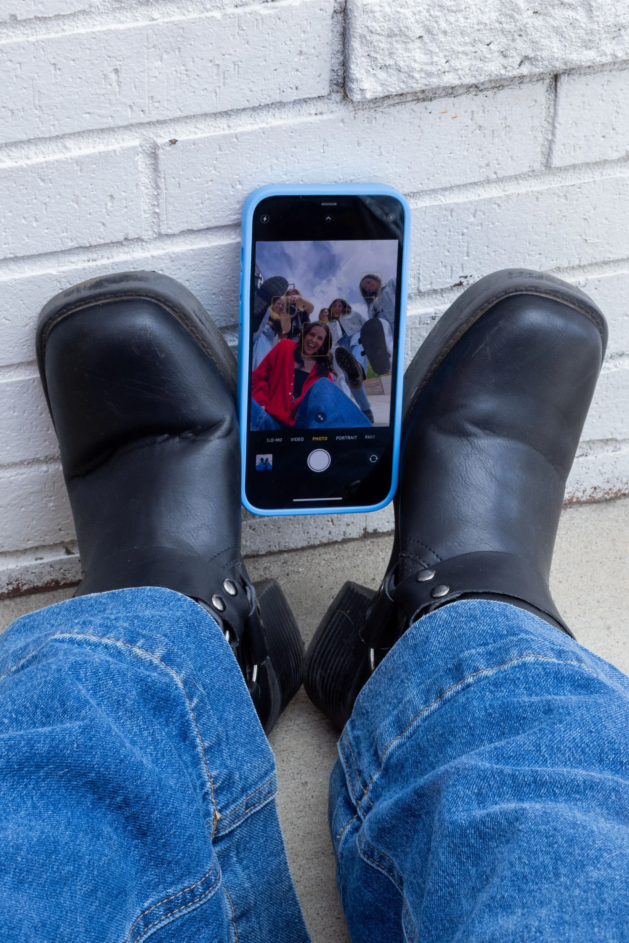 Black boots on the ground holding up a phone taking a selfie of six different girls against a white brick wall